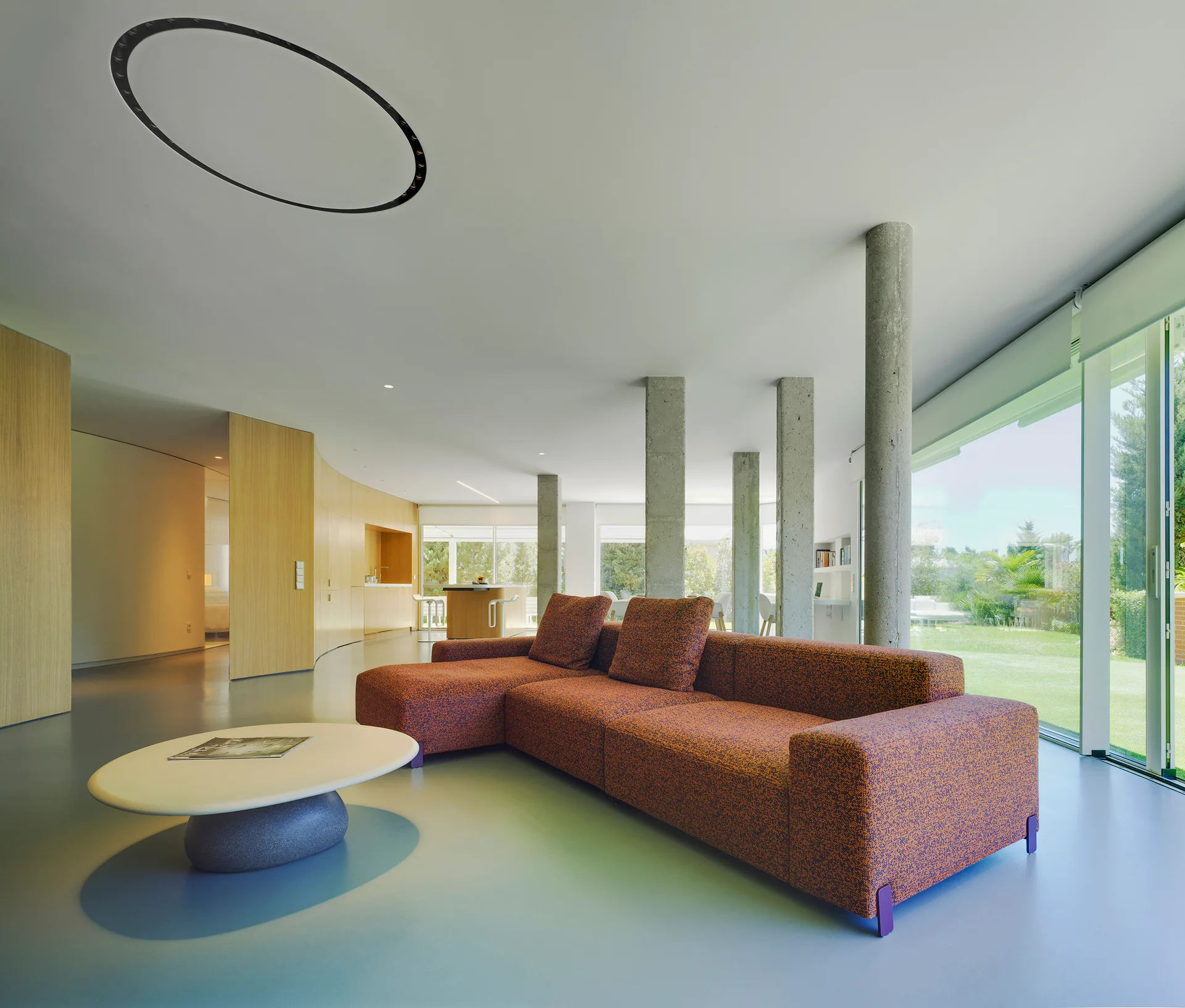 A modern living room with a large orange sectional sofa, a low oval coffee table, exposed concrete pillars, and floor-to-ceiling glass windows.