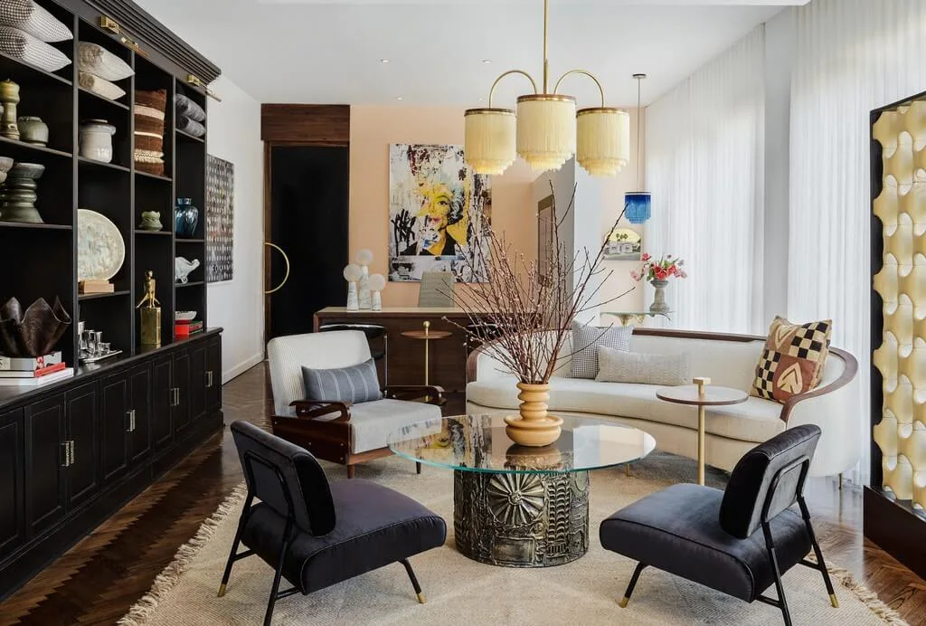 An eclectic living room features a curved beige sofa, a glass coffee table, two dark armchairs, and a large black built-in shelving unit.