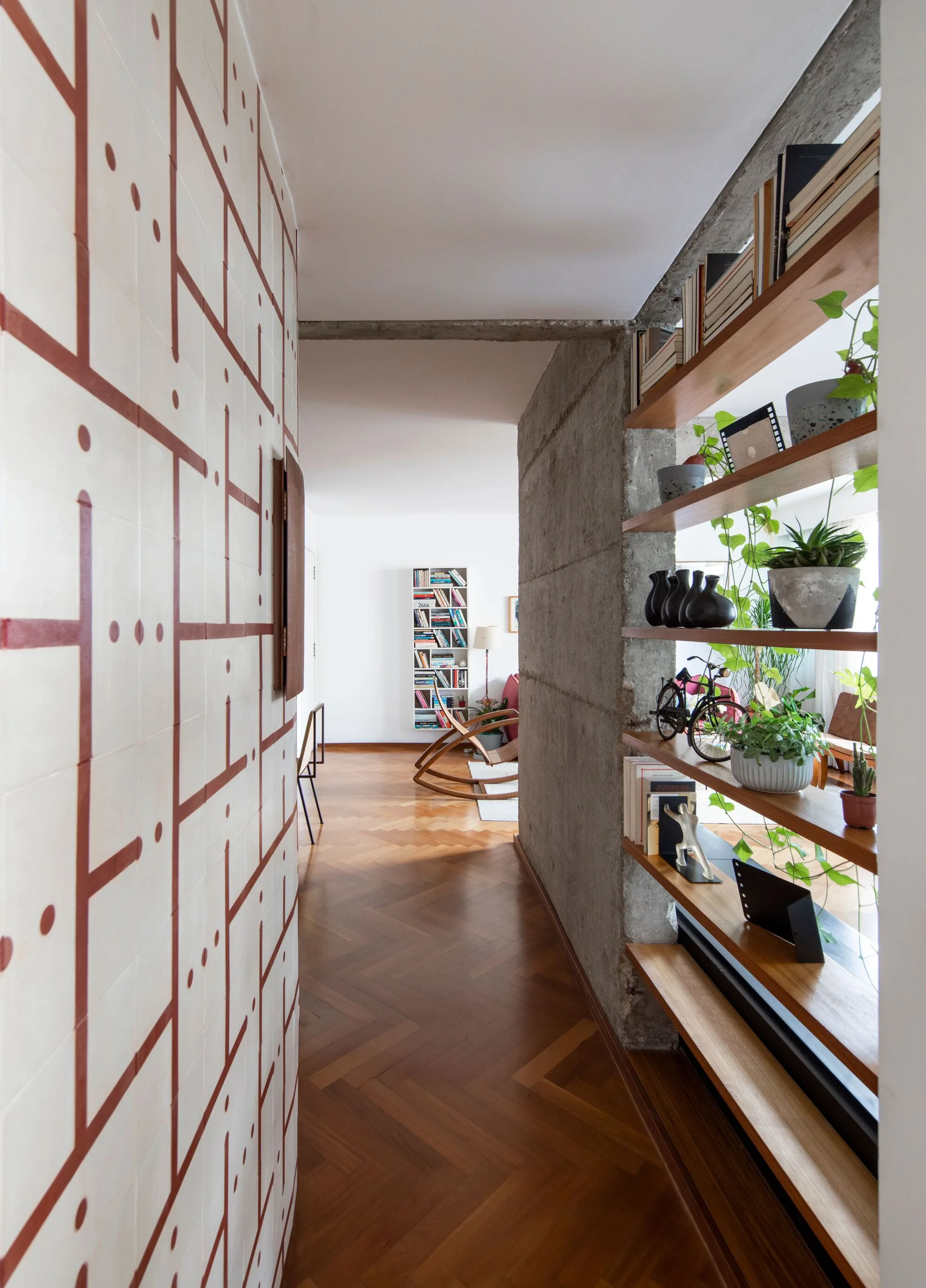 A residential hallway features a geometric patterned tile wall, a raw concrete partition with built-in wooden shelves, and herringbone wood flooring.