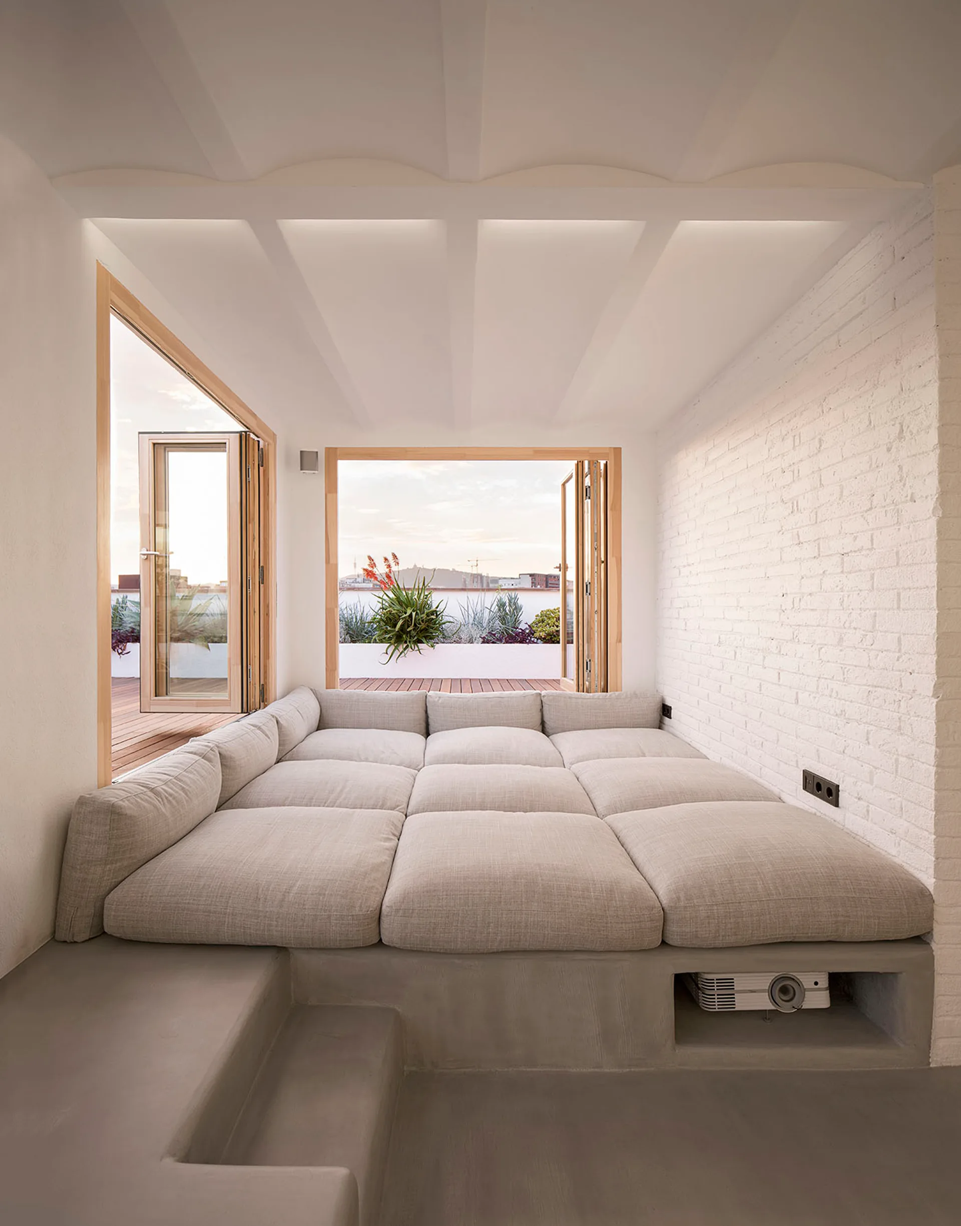 A minimalist indoor lounge features a large beige cushioned platform bed on a concrete base next to a white brick wall and open bi-fold doors.