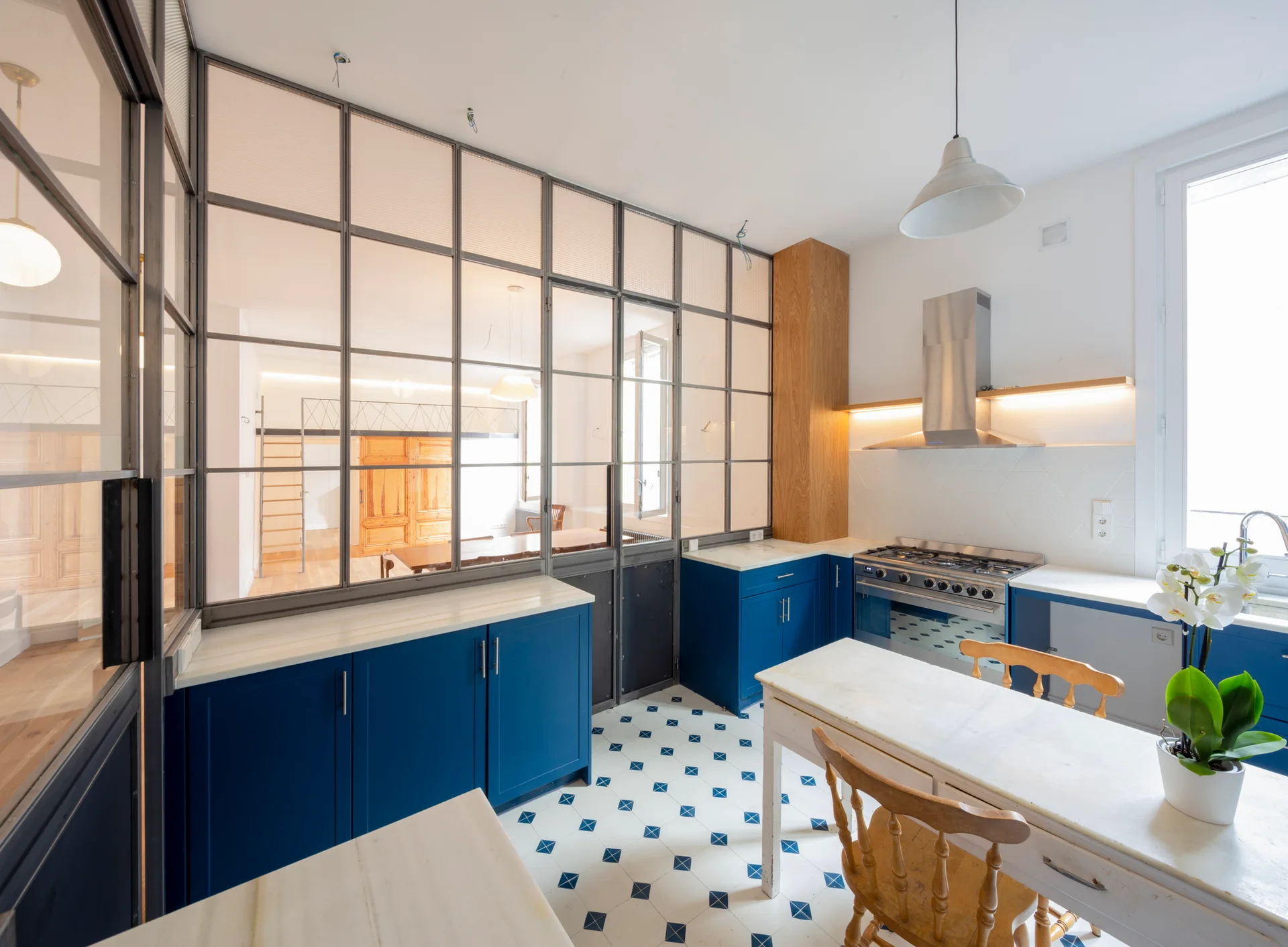 A bright kitchen featuring blue lower cabinetry, a patterned tile floor, and a large steel-framed glass partition wall.