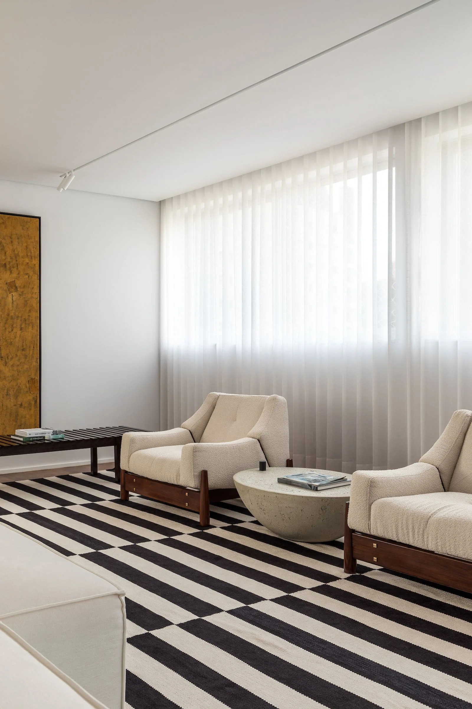 Two cream boucle armchairs sit on a bold black and white striped rug next to a low stone coffee table in a sunlit minimalist living room.