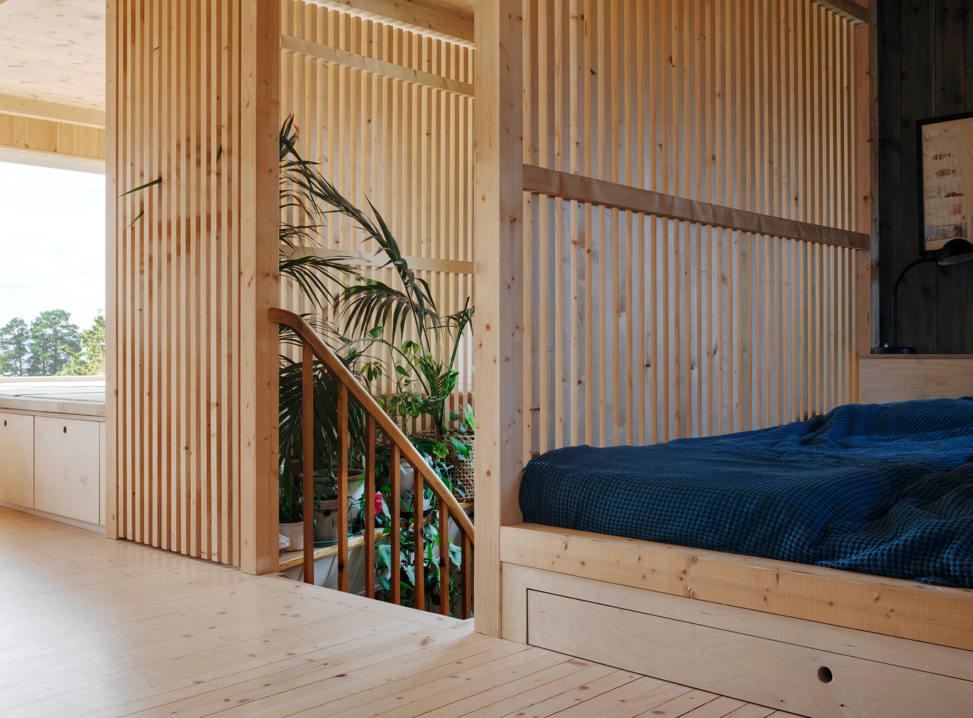 A minimalist light wood bedroom features a platform bed with dark blue bedding and a vertical slat partition wall next to a staircase.