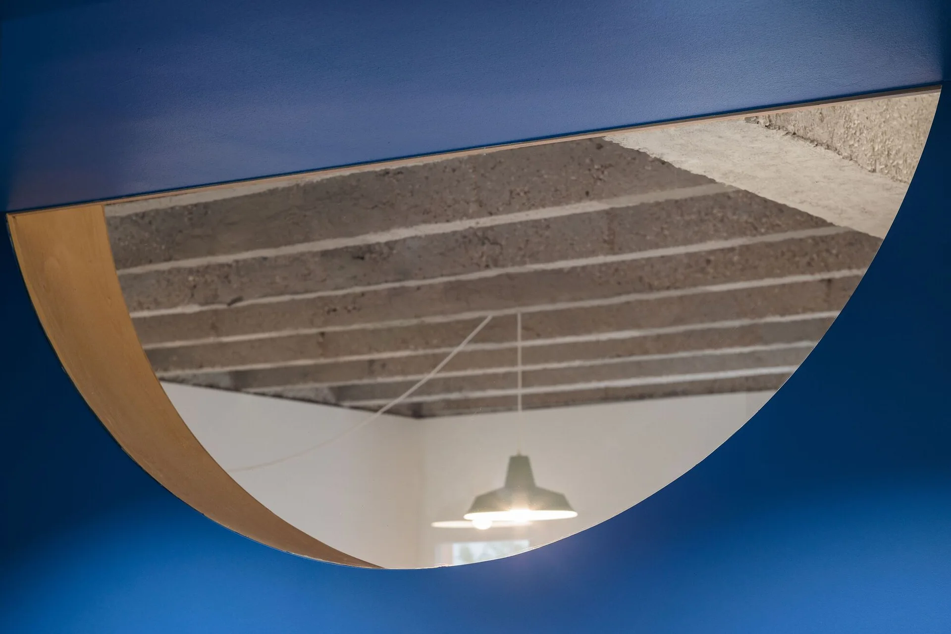 A semicircular wall mirror with wood trim is mounted on a deep blue wall, reflecting a textured concrete ceiling and a pendant light.
