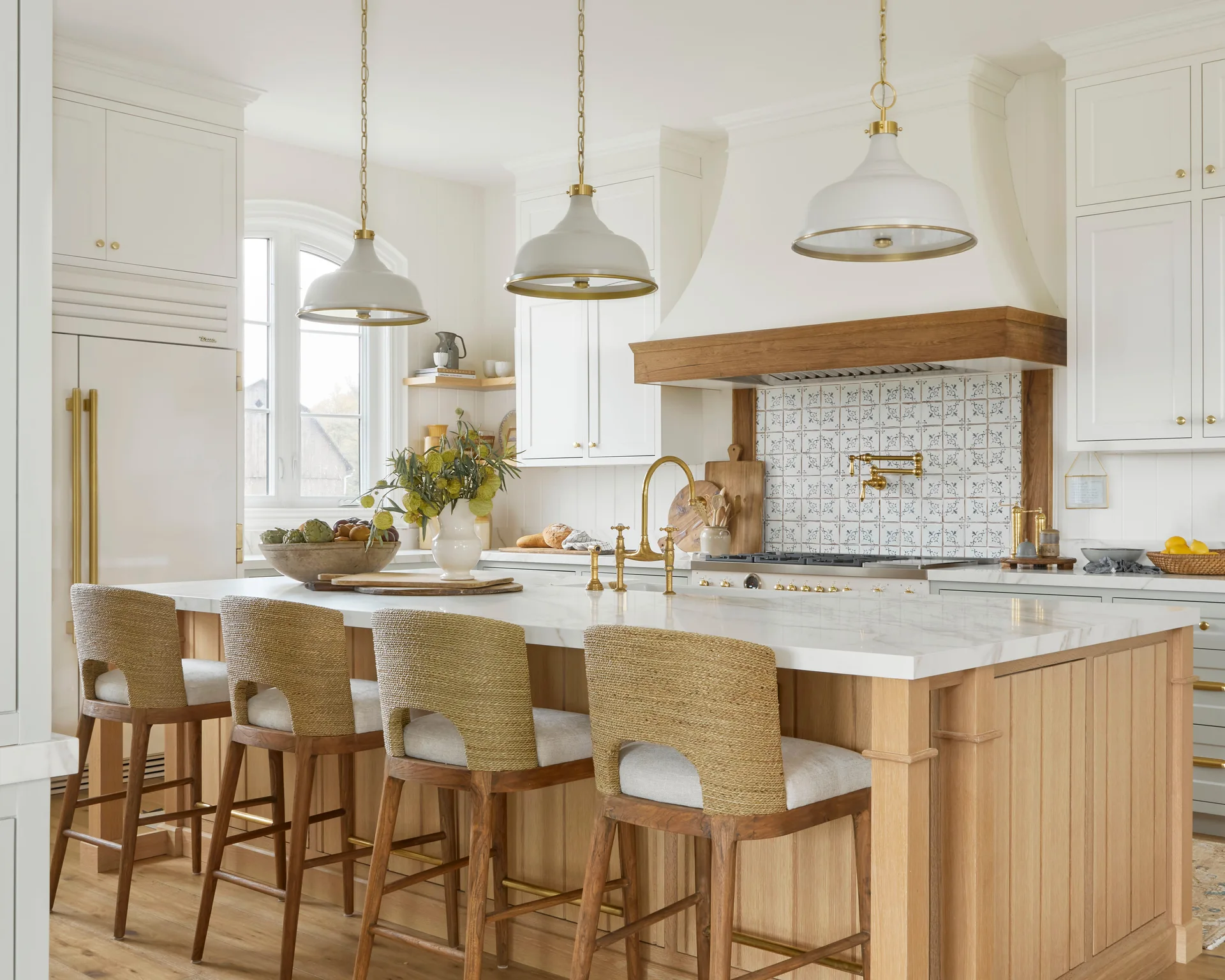 A bright kitchen features a light wood island with a marble countertop, four woven bar stools, and white dome pendant lights overhead.