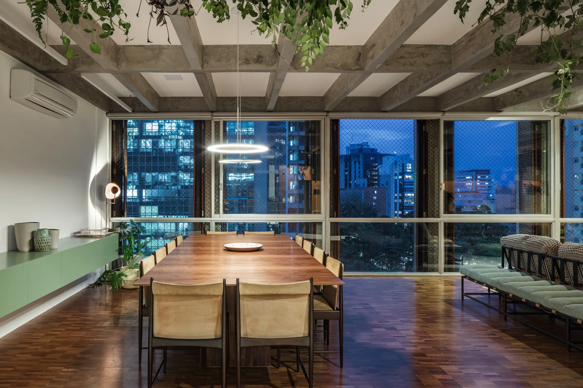 A modern dining room features a long wooden table with chairs under an exposed concrete grid ceiling with hanging plants and floor-to-ceiling windows.