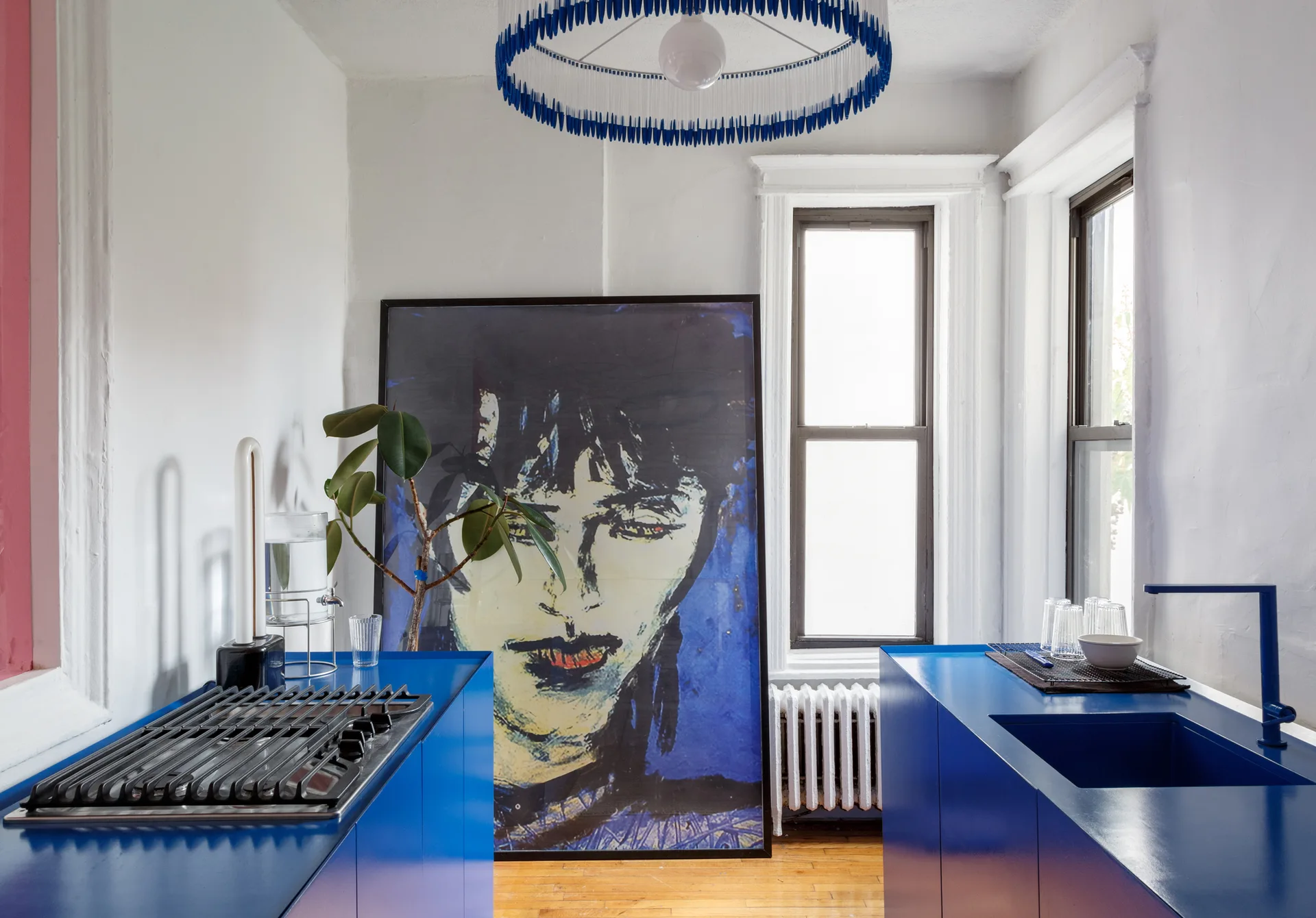 A galley kitchen features bright blue cabinets, a blue faucet, a large framed portrait leaning against the white wall, and a fringe pendant light.