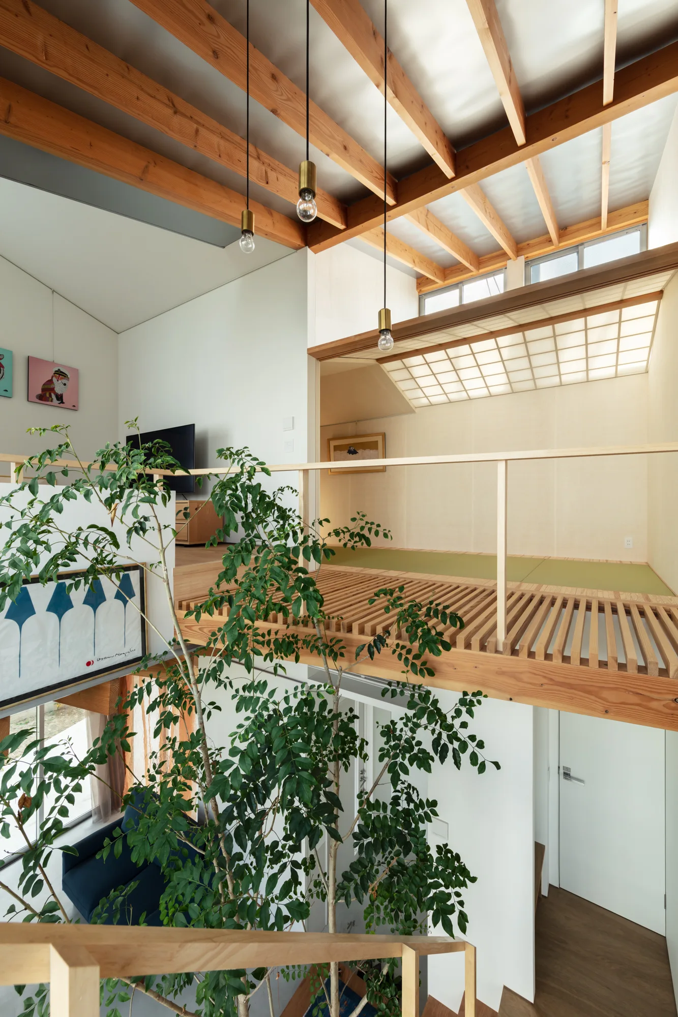 A modern split-level interior with exposed wooden ceiling beams, a leafy indoor tree, and a mezzanine level featuring green tatami mats.