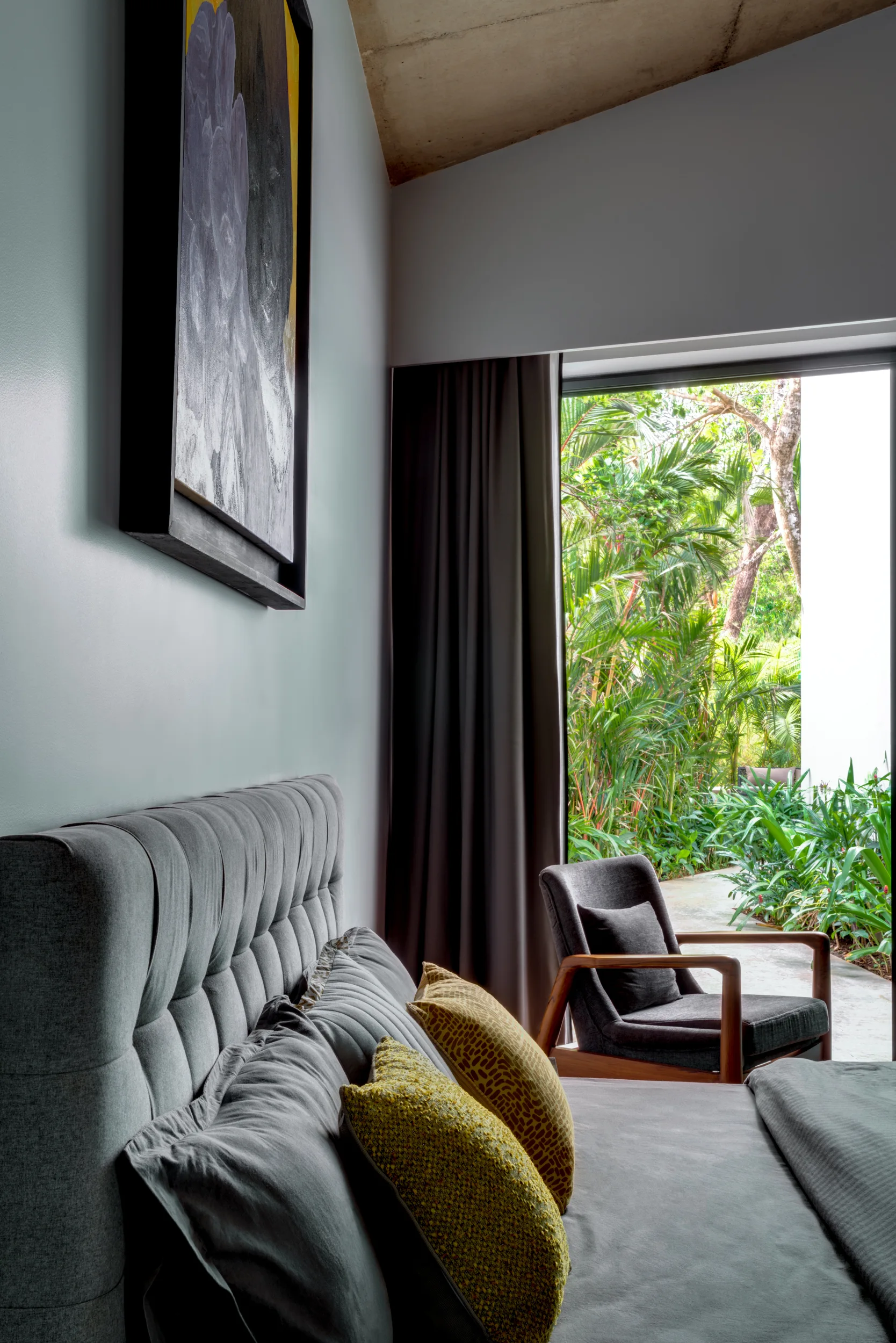 A grey tufted headboard bed and a wooden armchair face a large window overlooking a lush green garden under an angled concrete ceiling.