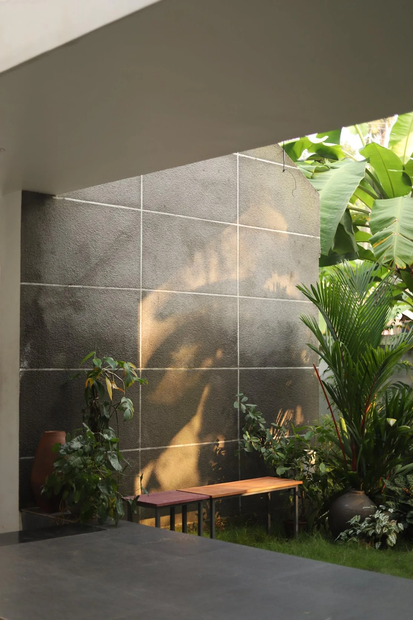 A modern outdoor patio features a large grey grid-tiled accent wall, tropical green plants, and a simple wooden bench on a grassy lawn.