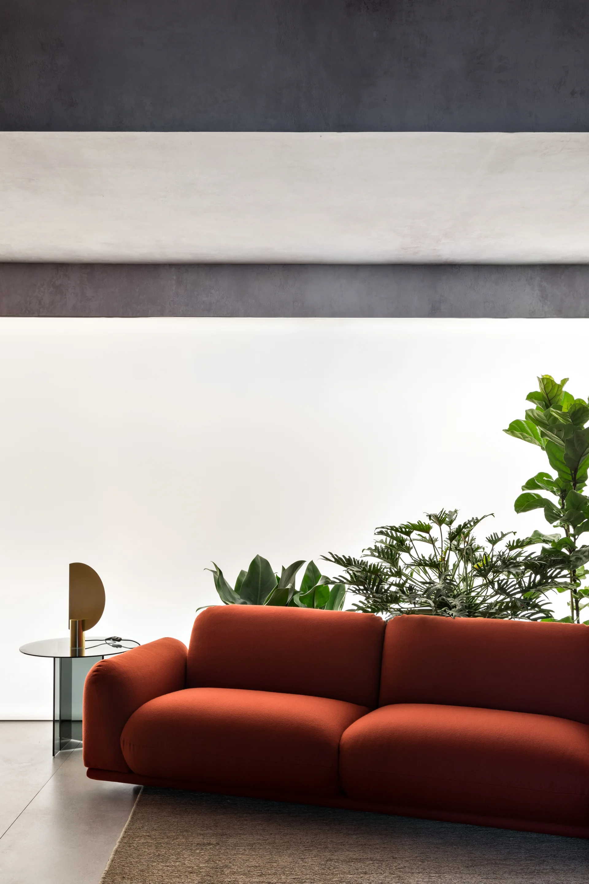A rust orange sofa sits in a minimalist living room featuring a glowing backlit wall, indoor plants, and a gold semi-circular table lamp.