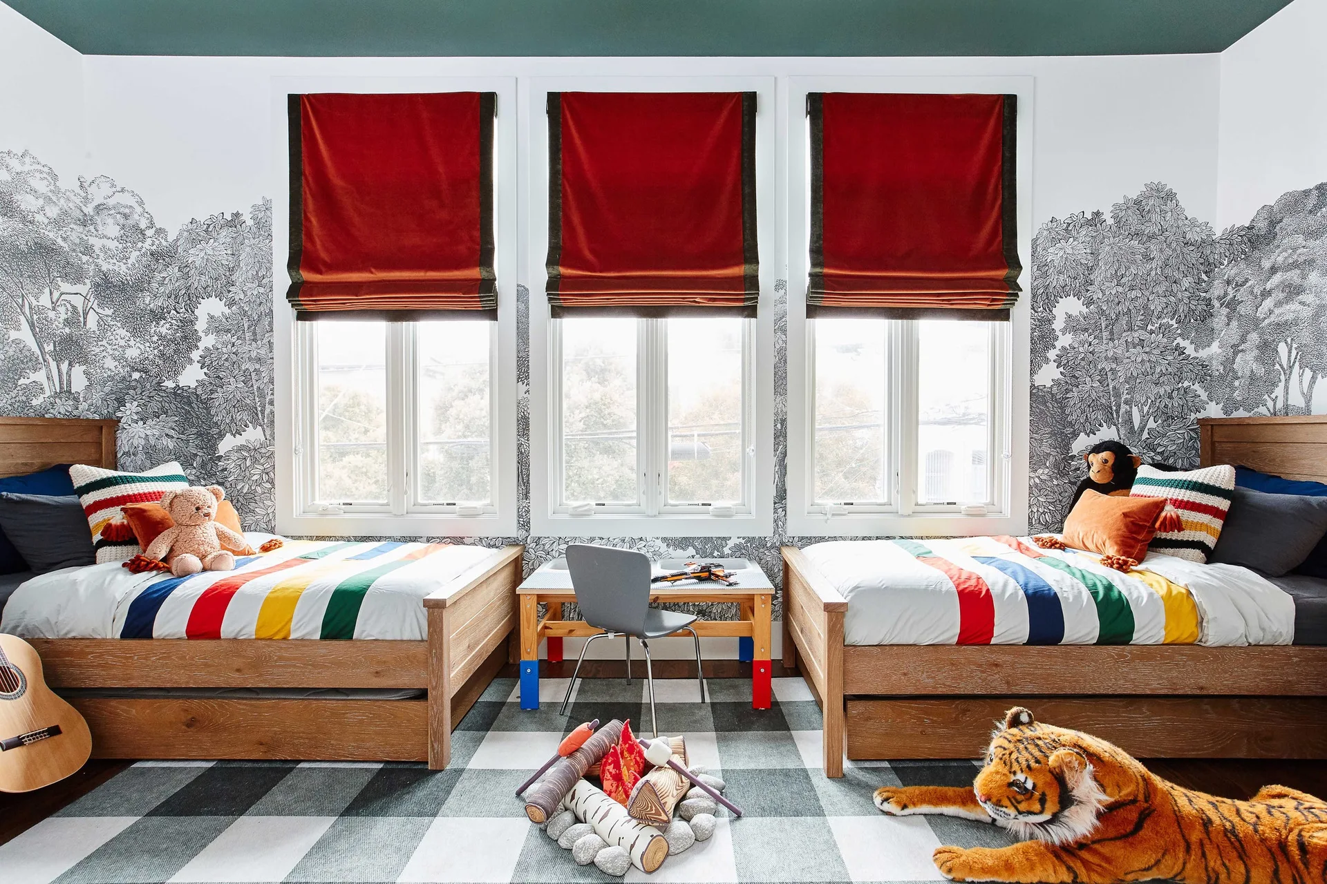 A children's bedroom features two wooden twin beds, black and white forest wallpaper, red roman shades, and a large checkered rug.