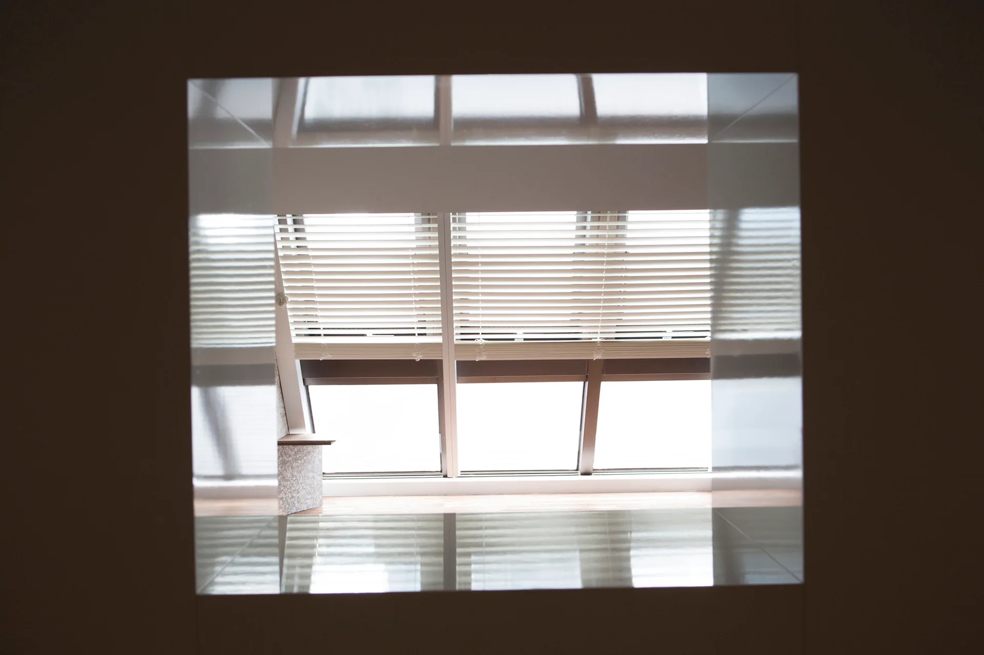 A view through a dark square opening reveals an angled skylight window with white slatted blinds and light wood flooring below.