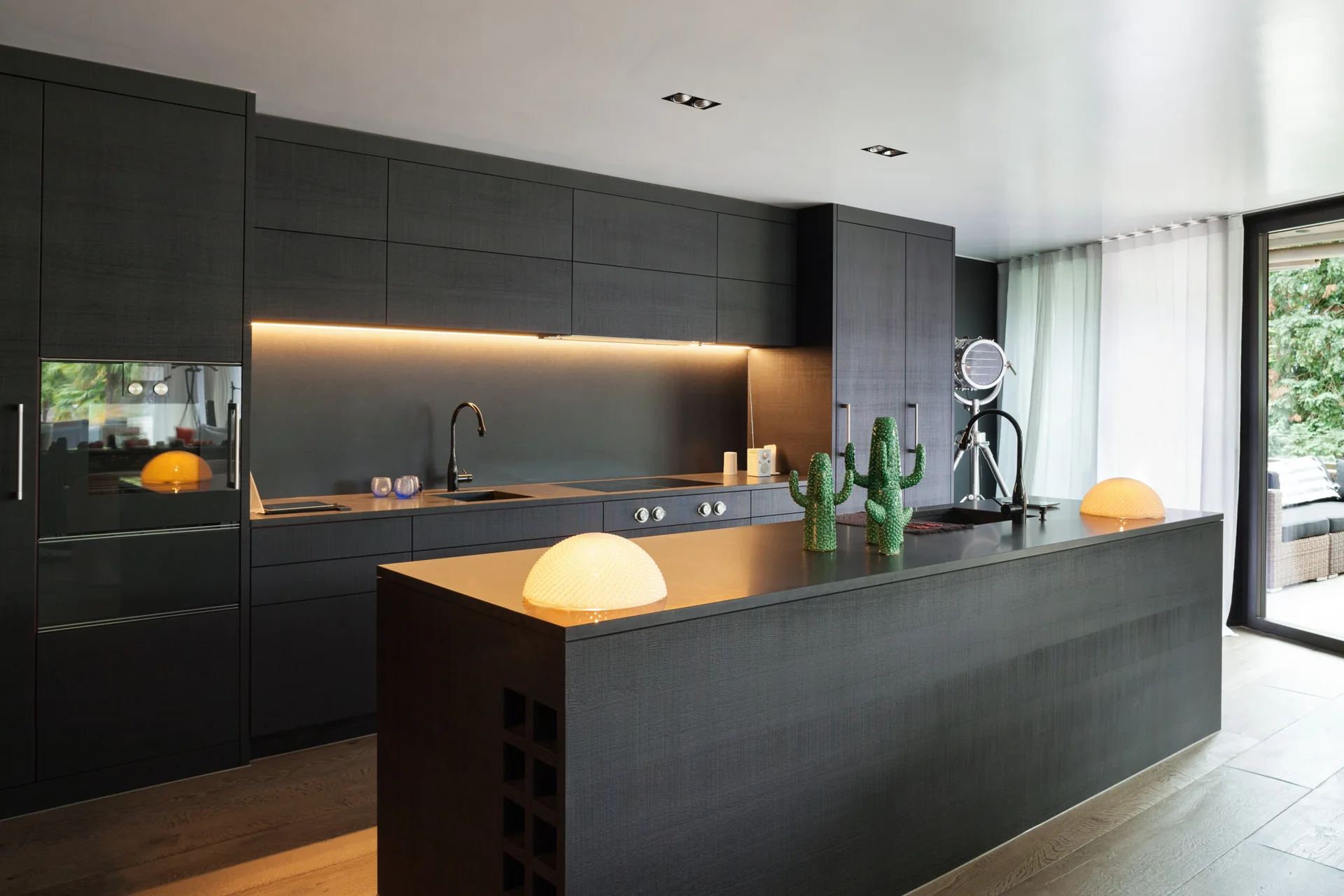 A modern black kitchen features dark wood-grained cabinets, a large central island, warm under-cabinet strip lighting, and two dome lamps on the counter.