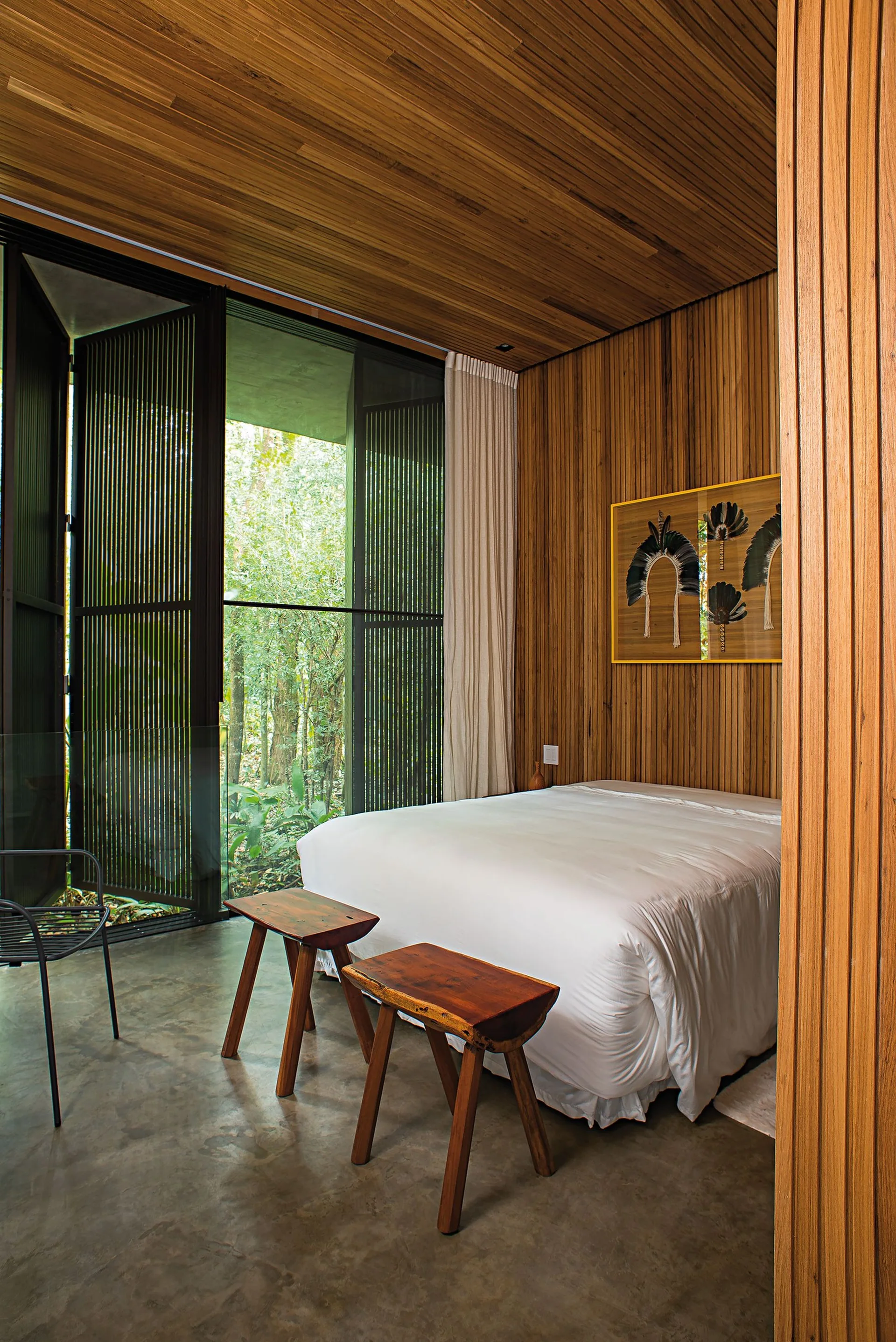 A wood-paneled bedroom with a white bed, two wooden stools on a concrete floor, and floor-to-ceiling glass windows showcasing a lush green forest view.
