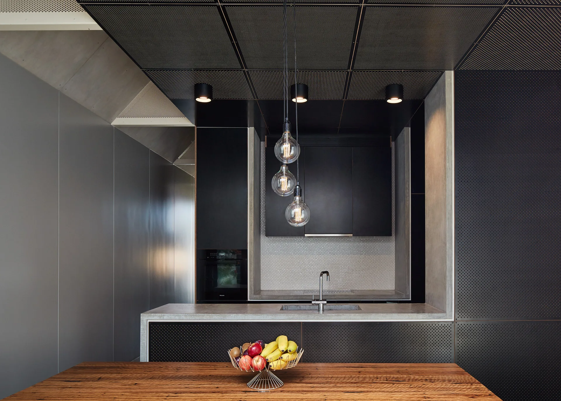 A modern industrial kitchen with a concrete island, black cabinetry, perforated metal wall and ceiling panels, and three glass globe pendant lights.