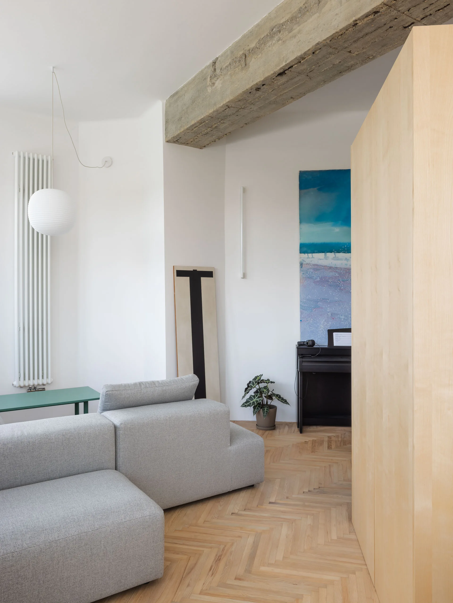 A light grey modular sofa sits on herringbone wood floors beneath an exposed concrete ceiling beam in a minimalist white living room.