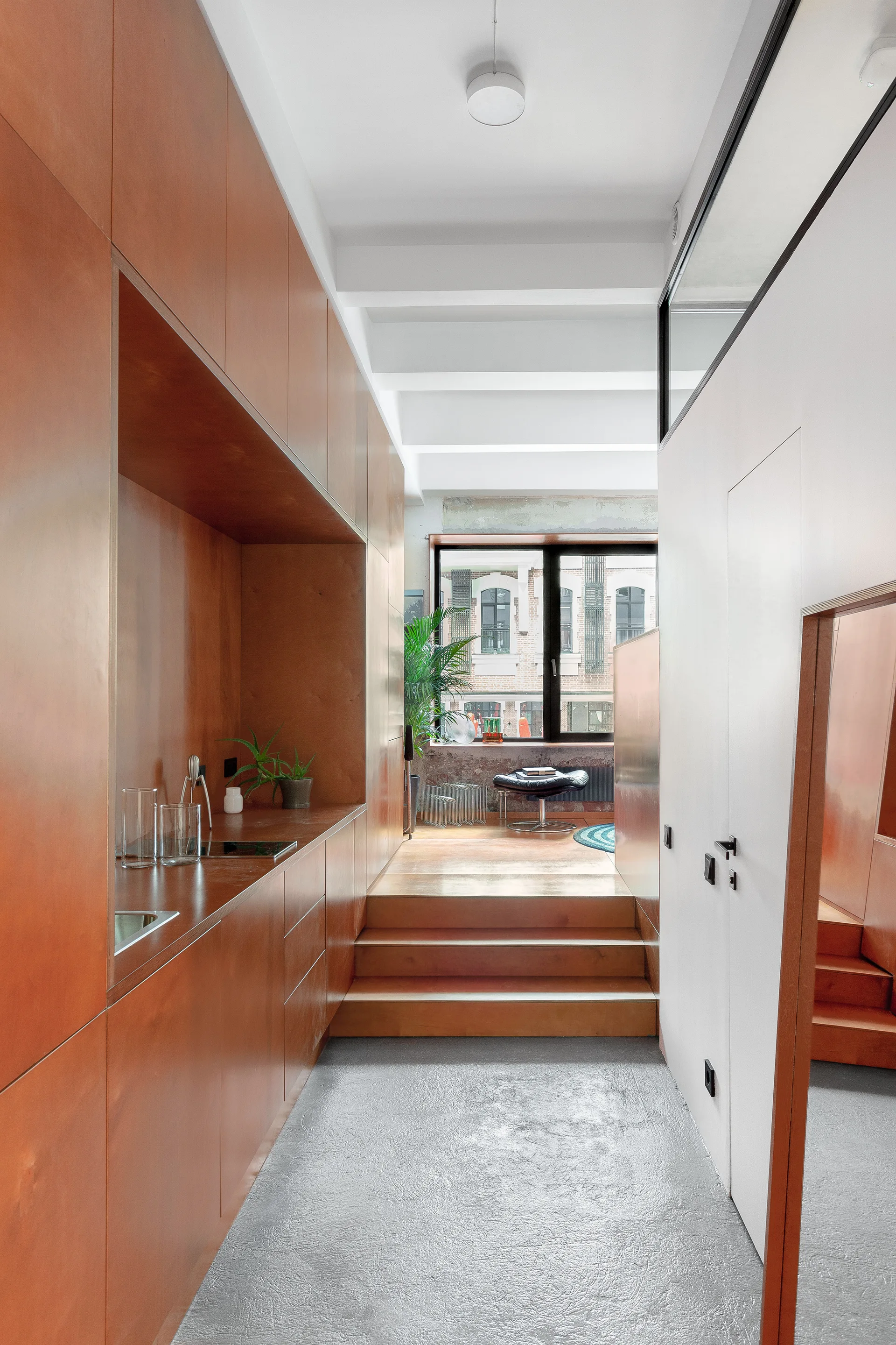 A split-level apartment interior featuring a large warm wood wall with built-in kitchen cabinetry, concrete flooring, and steps leading to a raised lounge.