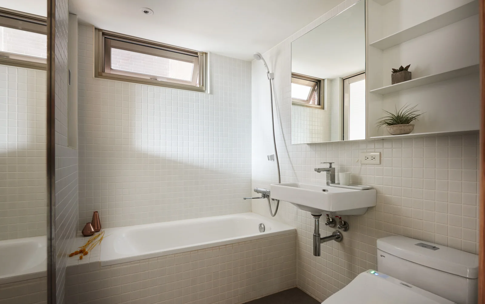A compact bathroom features white square-tiled walls, a built-in bathtub, a wall-mounted sink, and a high horizontal transom window.