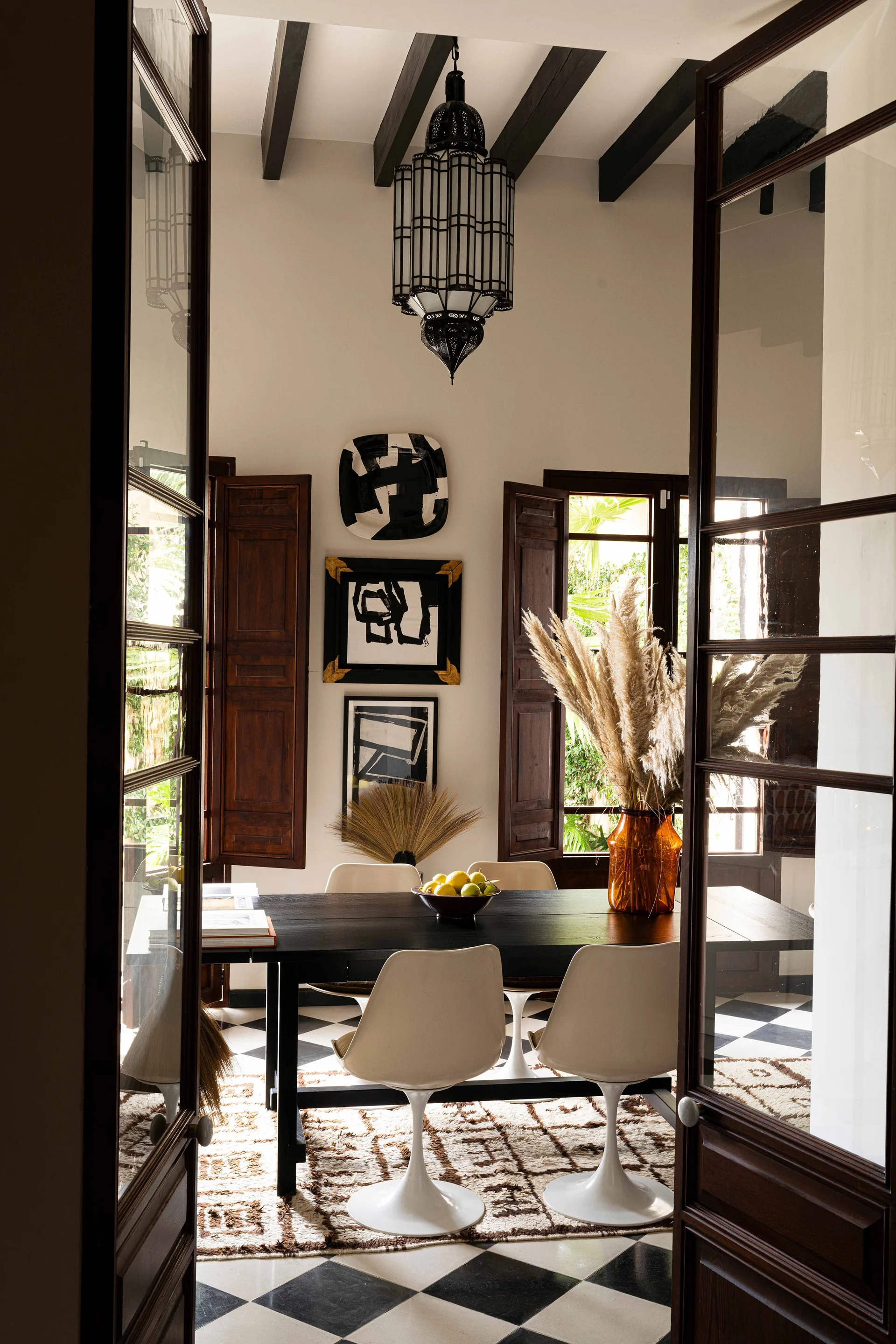 A dining room with a black table, white tulip chairs, and dark wood ceiling beams, viewed through open glass-paned doors.