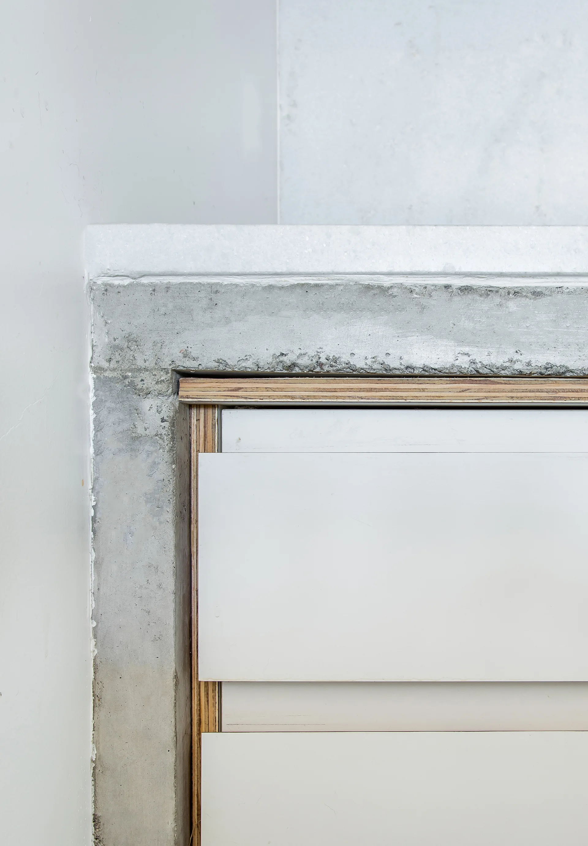 A close-up view of a minimalist cabinet with exposed plywood edges, surrounded by a raw concrete frame and a white stone countertop.