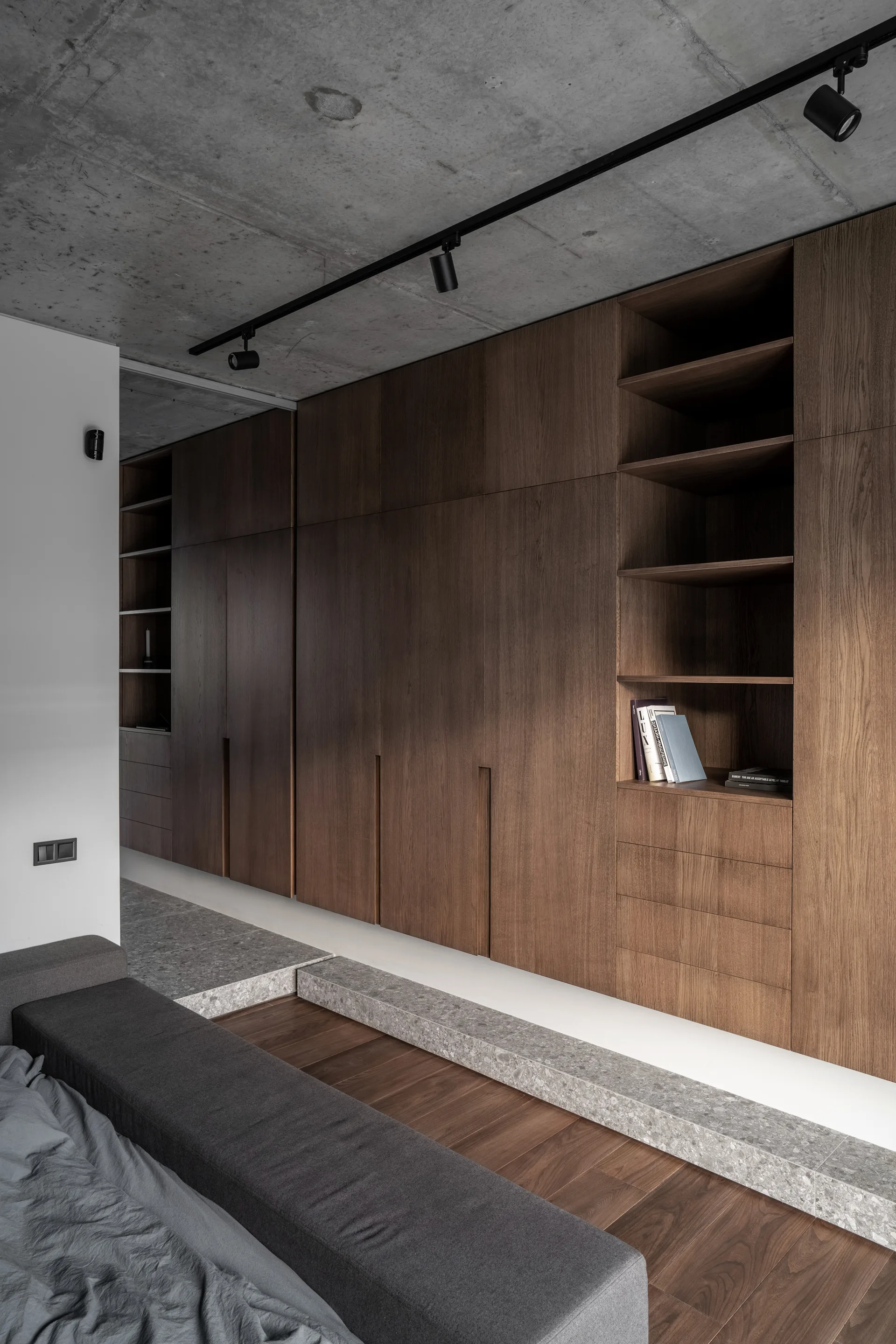 A modern minimalist bedroom features a large built-in wood wardrobe unit, a low-profile bed, an exposed concrete ceiling, and track lighting.