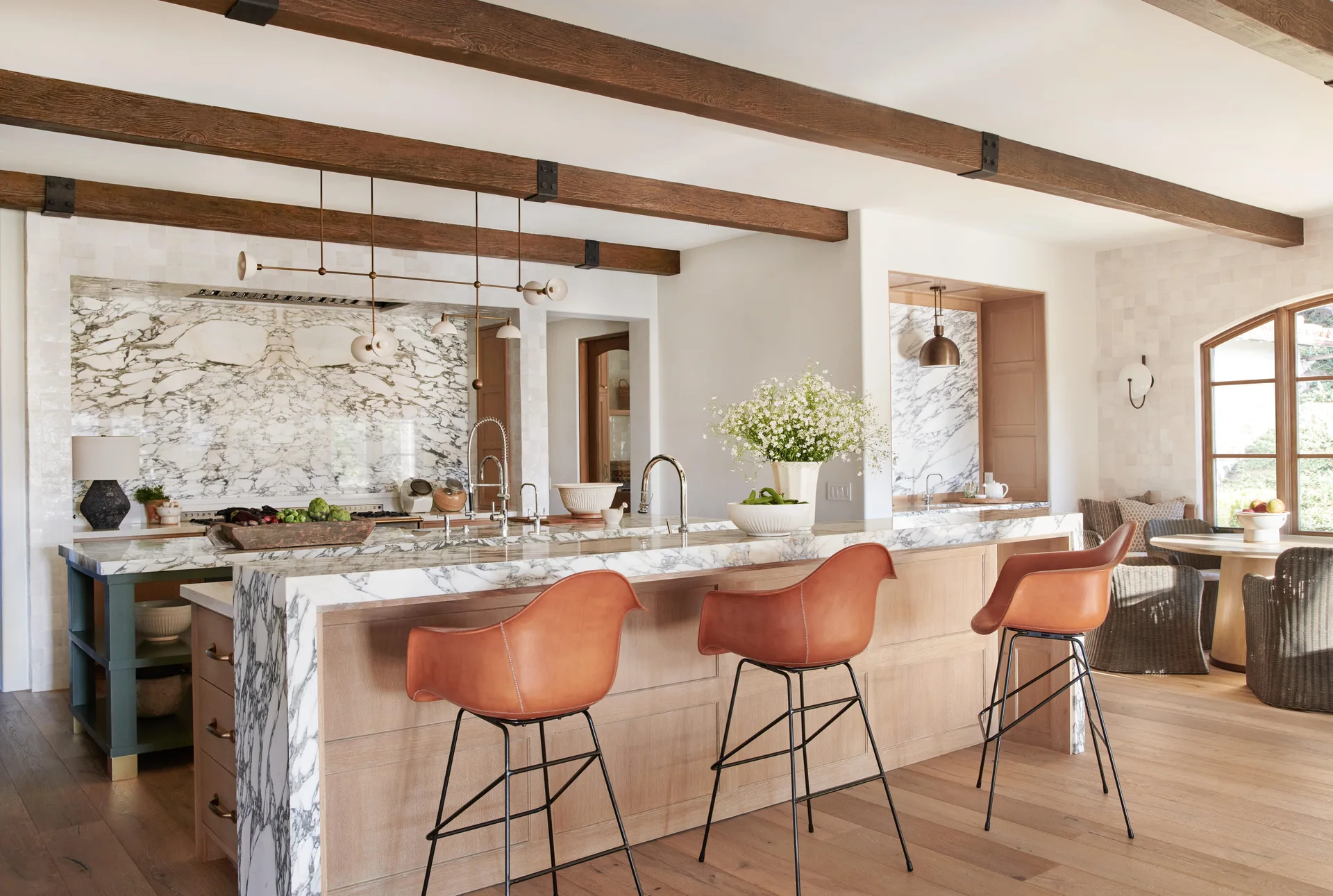 A spacious kitchen features a marble waterfall island with cognac leather stools, dark wood ceiling beams, and matching stone backsplash.