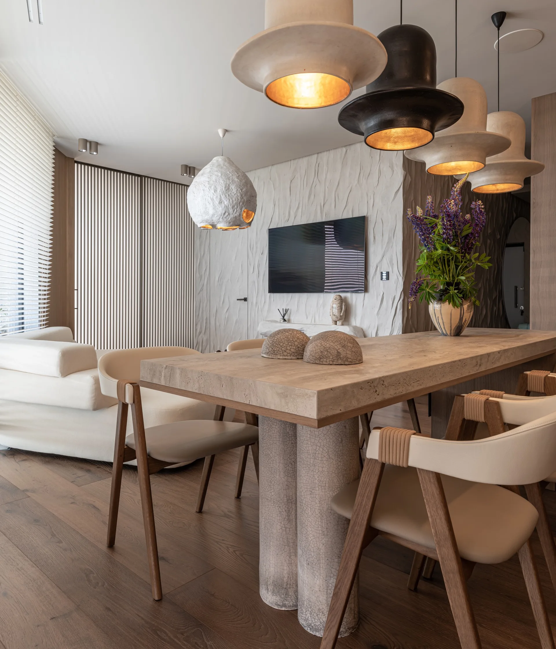 A contemporary dining space features a heavy stone table with wood and leather chairs, unique pendant lights, and a textured white accent wall.