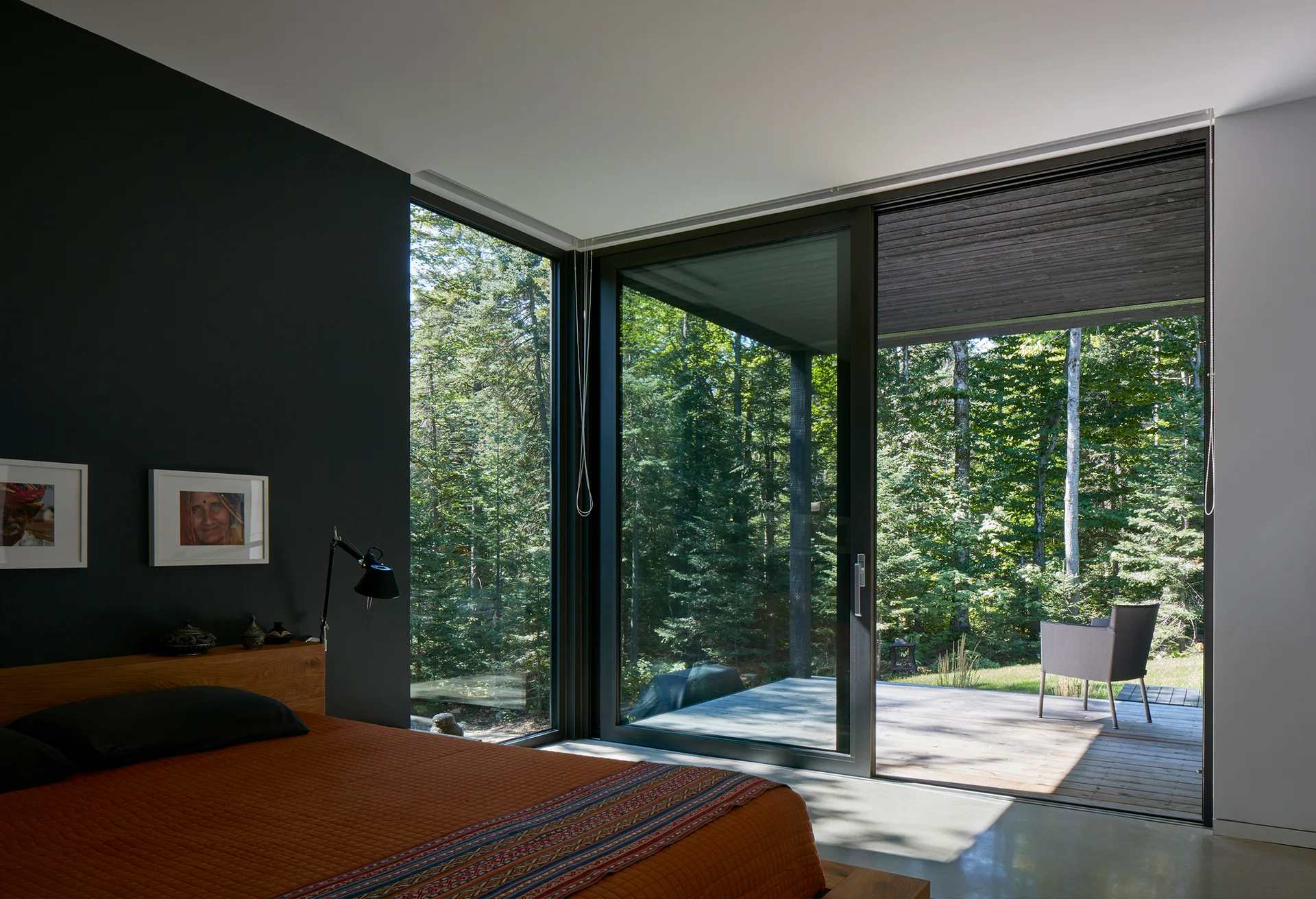 A modern bedroom features a low wooden platform bed against a dark charcoal wall next to large sliding glass doors opening onto a forest deck.