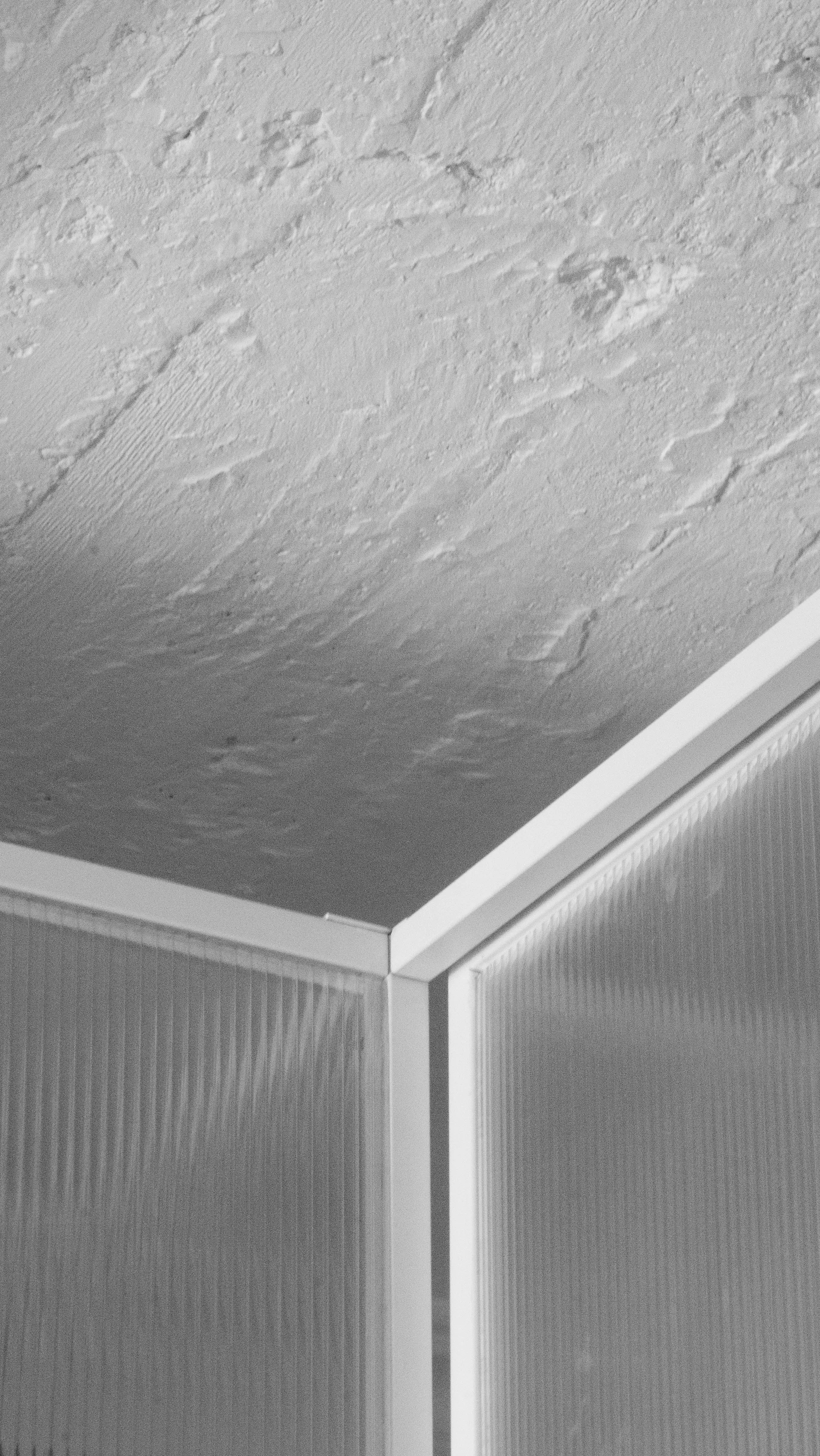 A close-up of a white textured plaster ceiling meeting a translucent ribbed glass partition wall framed in white metal.