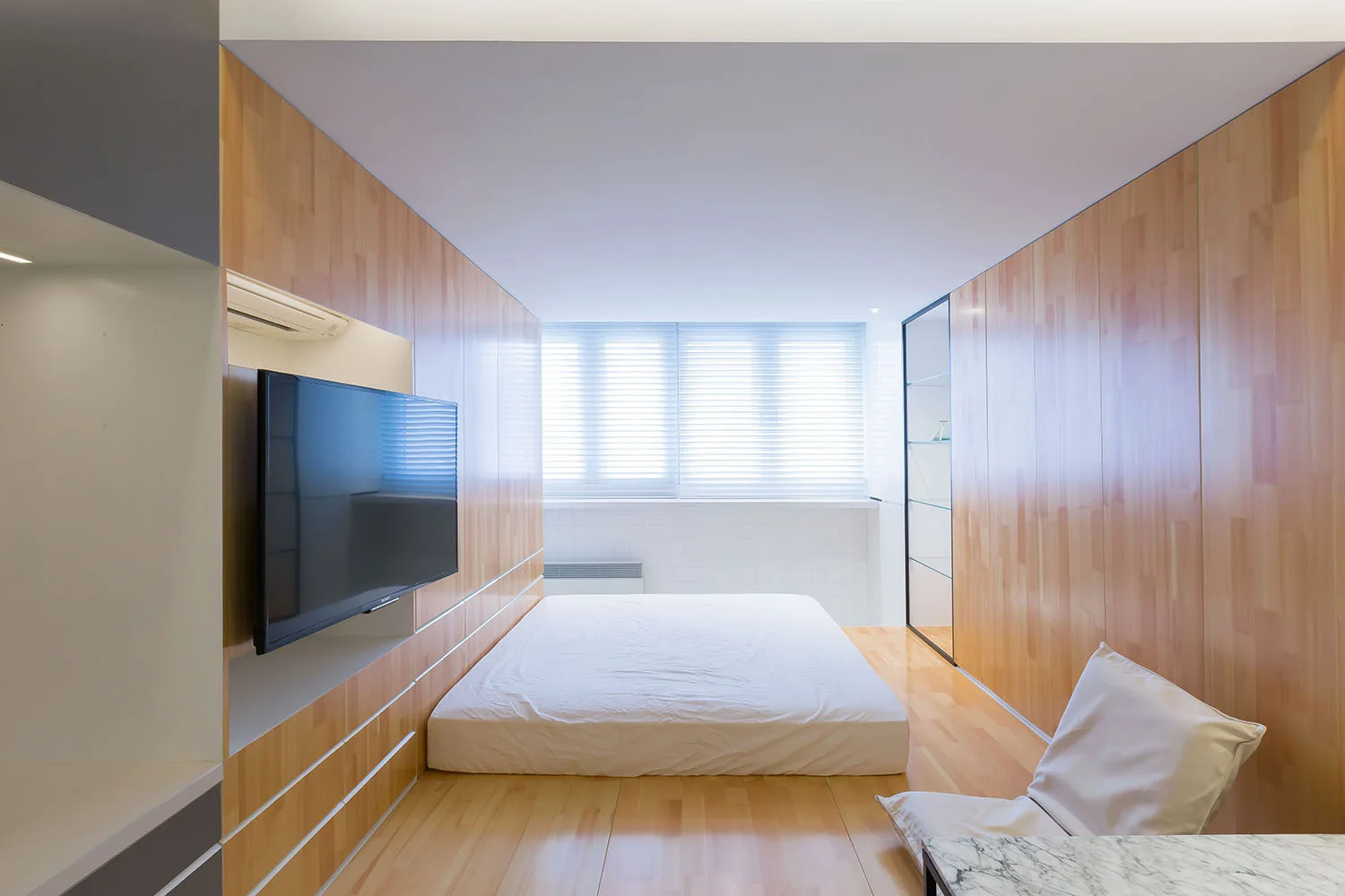 A minimalist bedroom with light wood paneling on the walls and floor, a low platform bed, a wall-mounted TV, and window blinds.