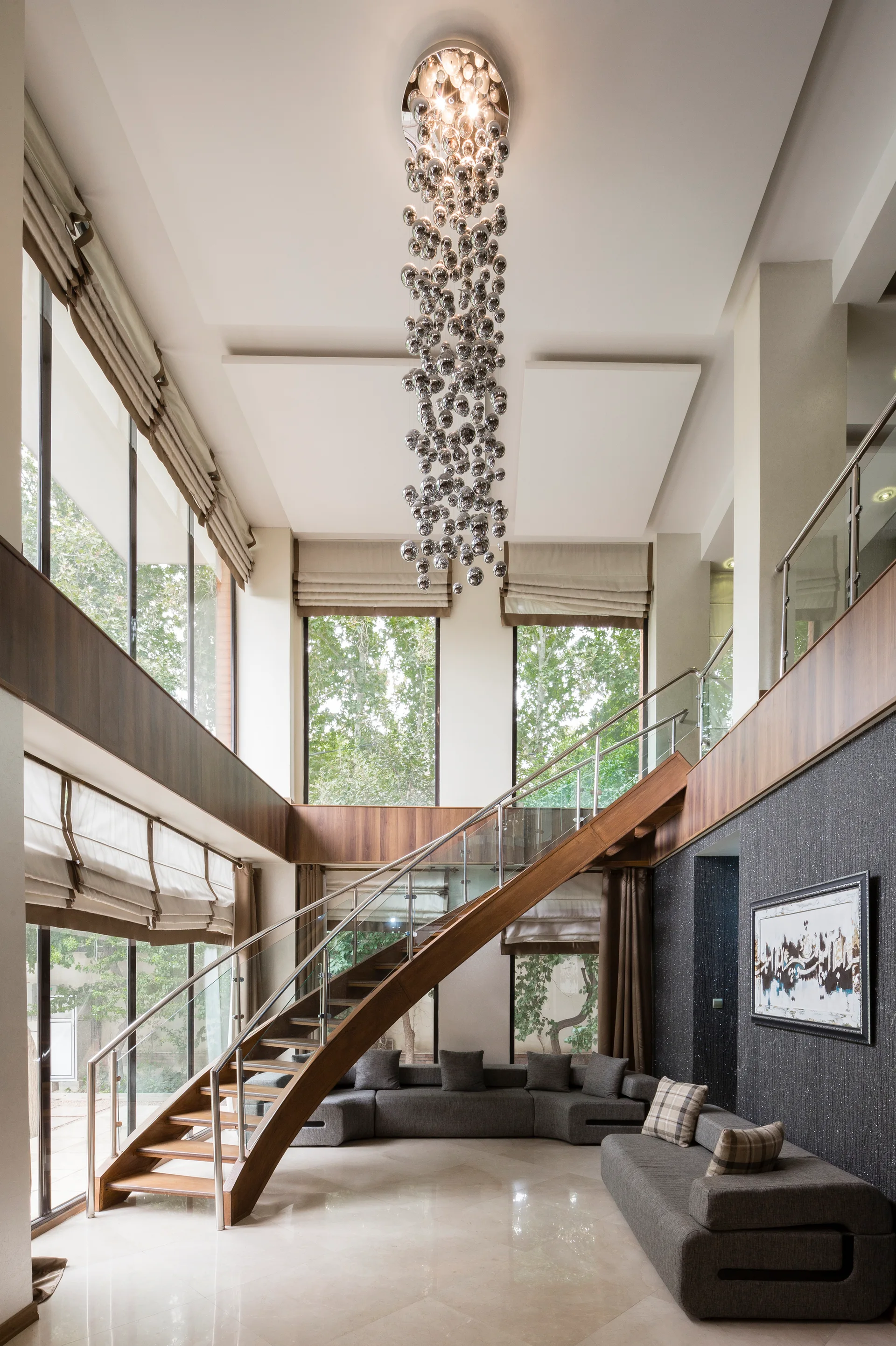 A modern double-height living room featuring a wooden staircase with glass railings, a grey sectional sofa, and a tall cascading chandelier.