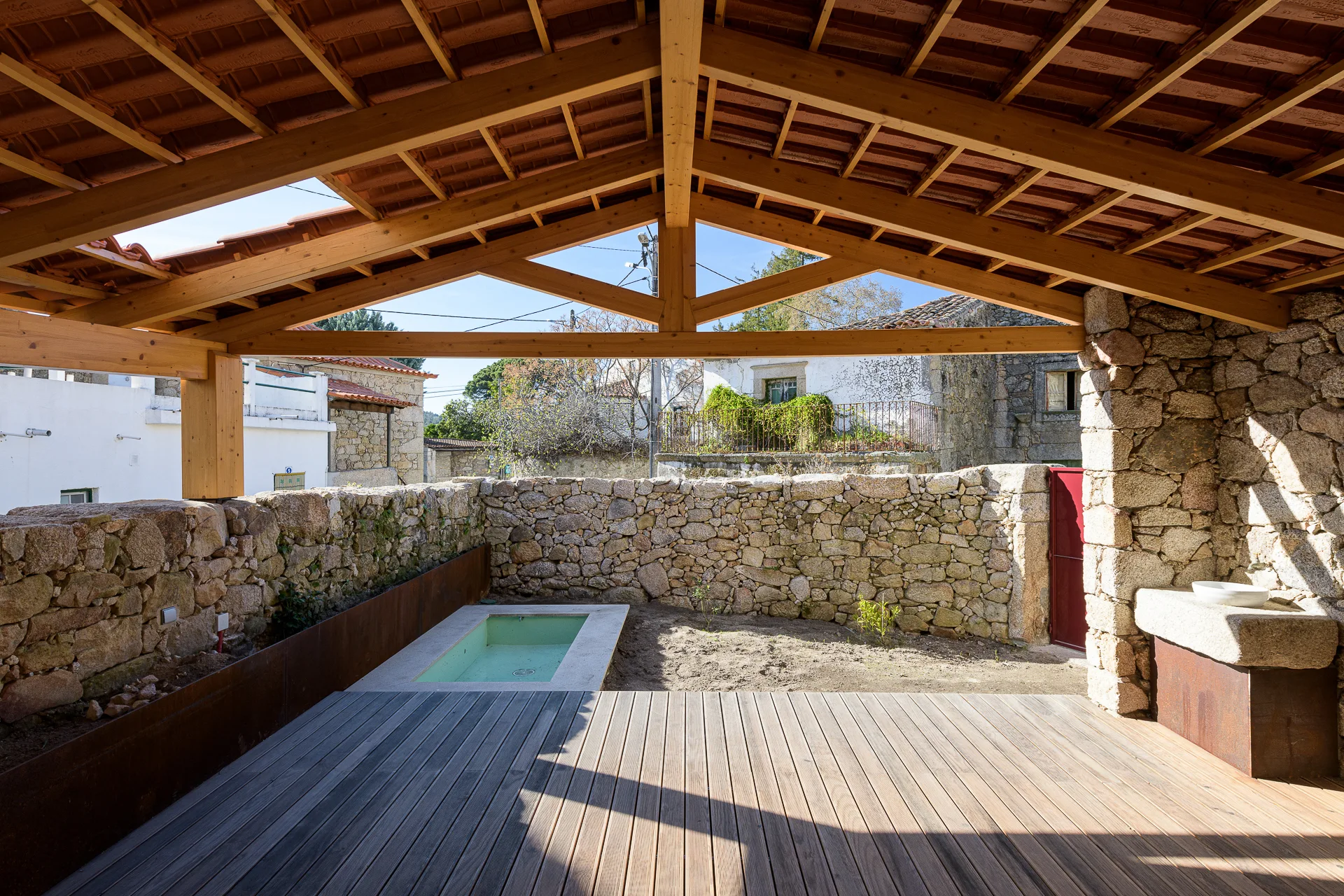 A covered wooden patio with plank flooring overlooks a sunken rectangular pool surrounded by rough stacked stone walls under a pitched timber roof.