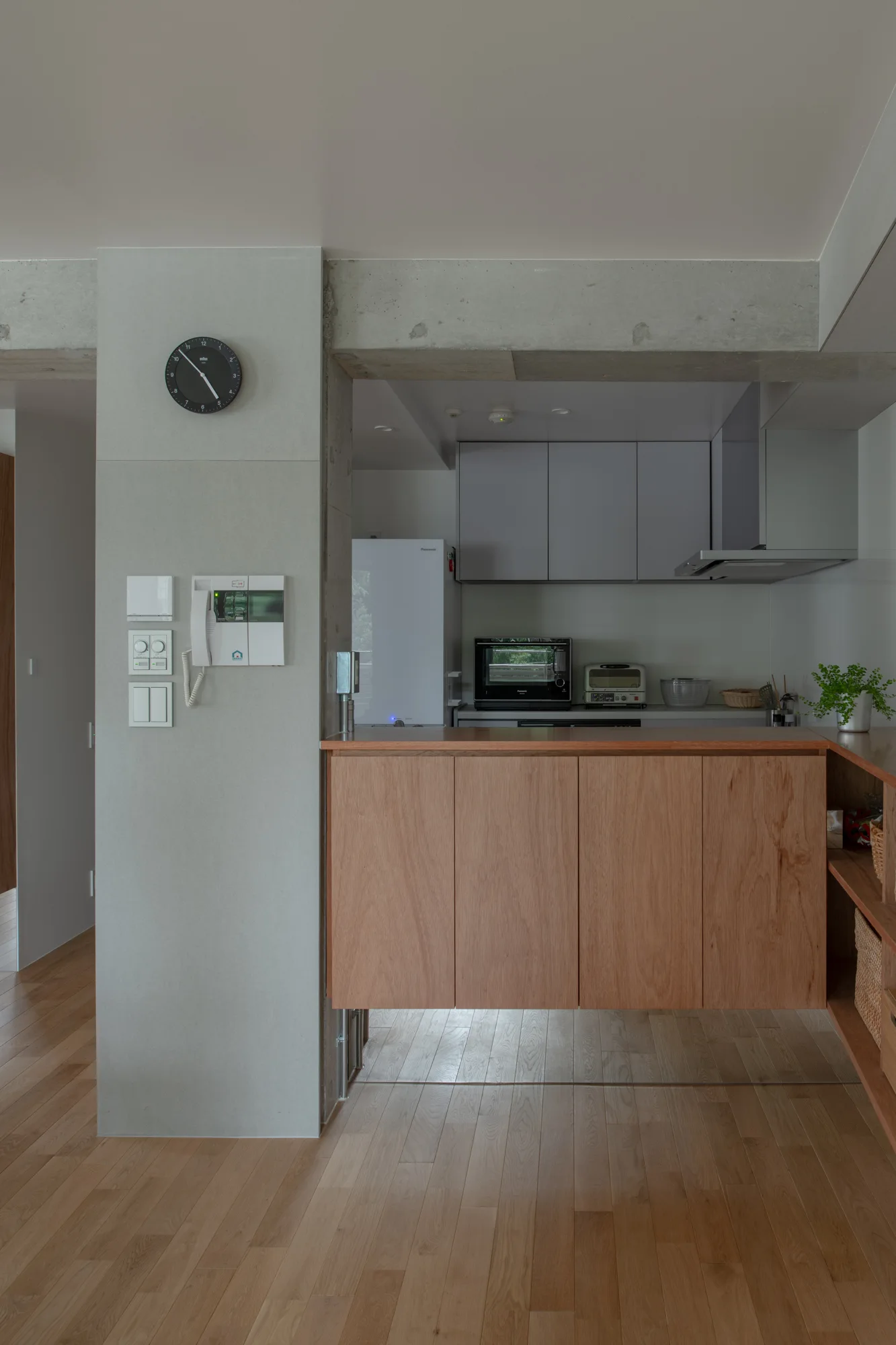 A modern residential kitchen features a grey concrete pillar beside a natural wood counter cabinet, with wood floors and grey upper cabinetry.