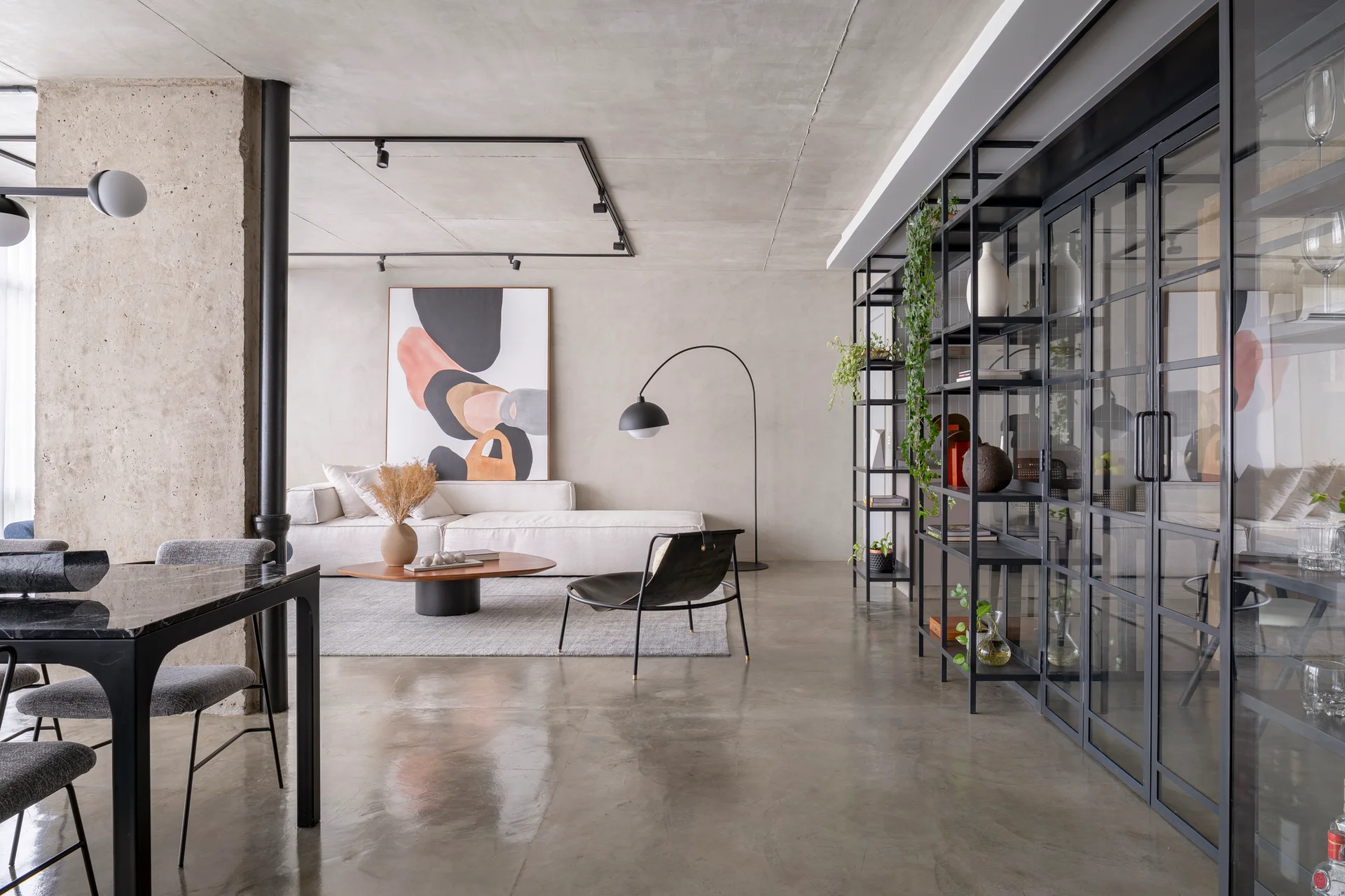 An open-plan living room features a white sectional sofa, raw concrete ceiling and column, and a large black metal display shelving unit.