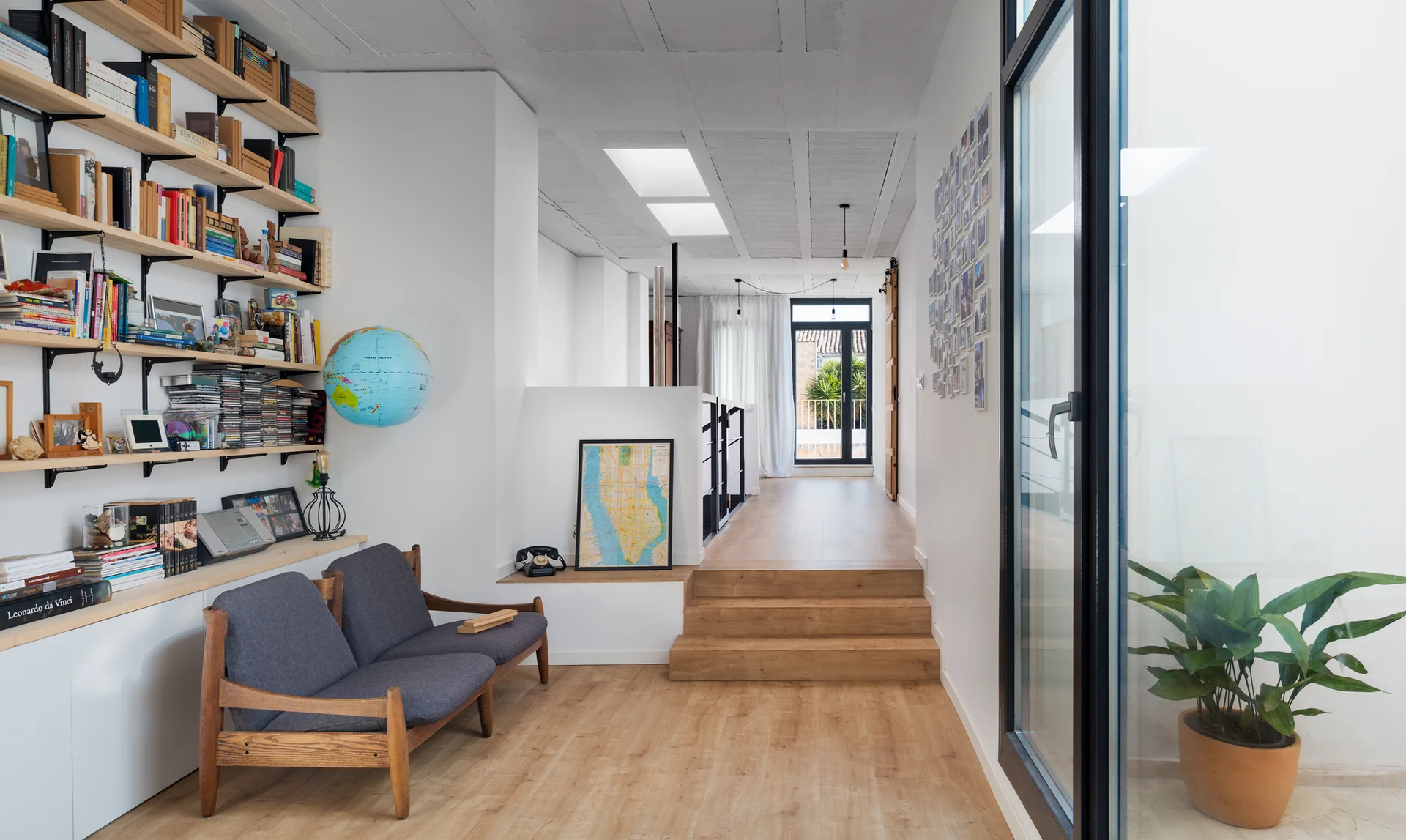 A bright hallway with wood flooring and tiered stairs features a gray wooden sofa, open bookshelves, and ceiling skylights.