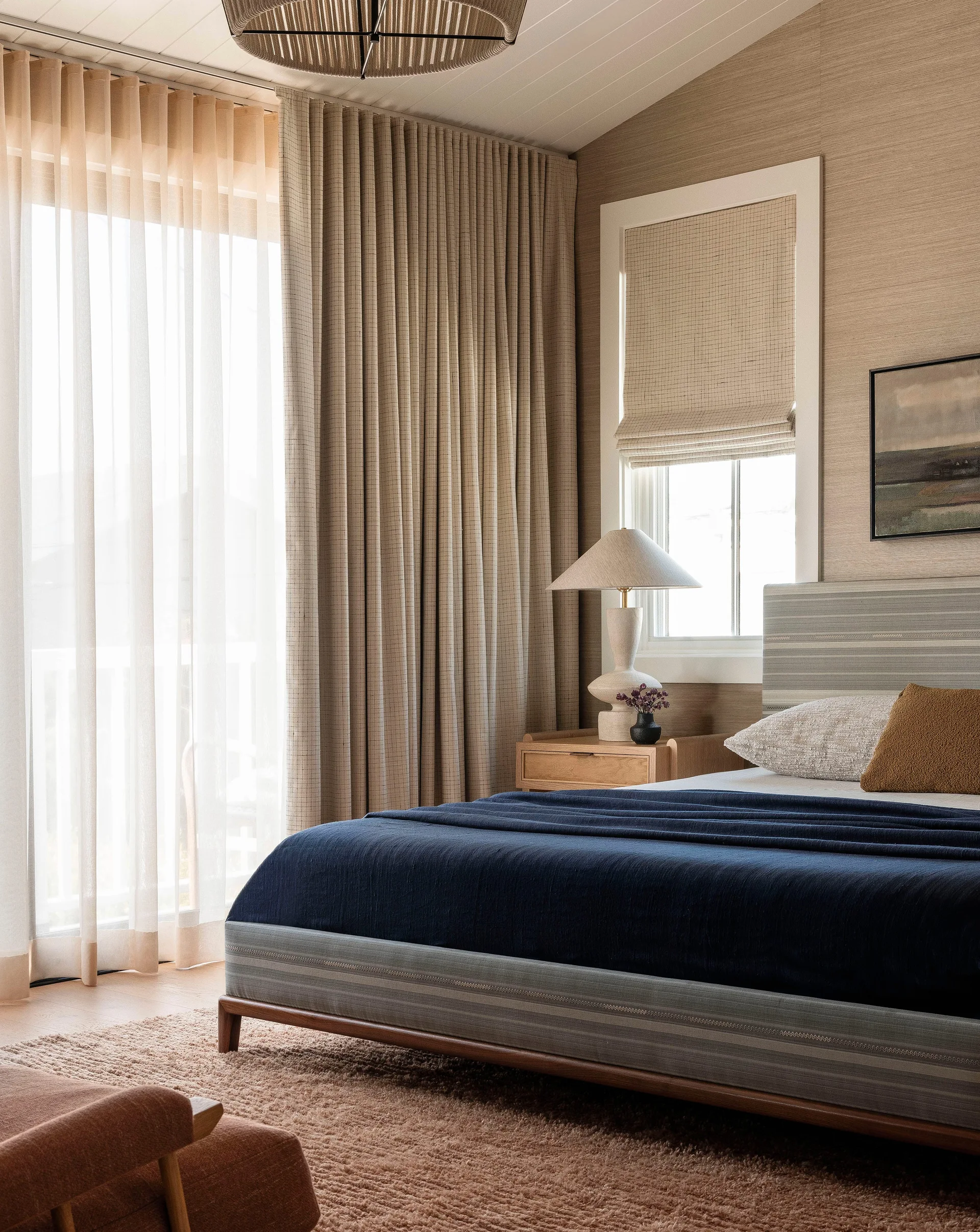 A bedroom features an upholstered striped bed with a navy blue blanket, layered beige curtains, a wooden nightstand, and a textured table lamp.