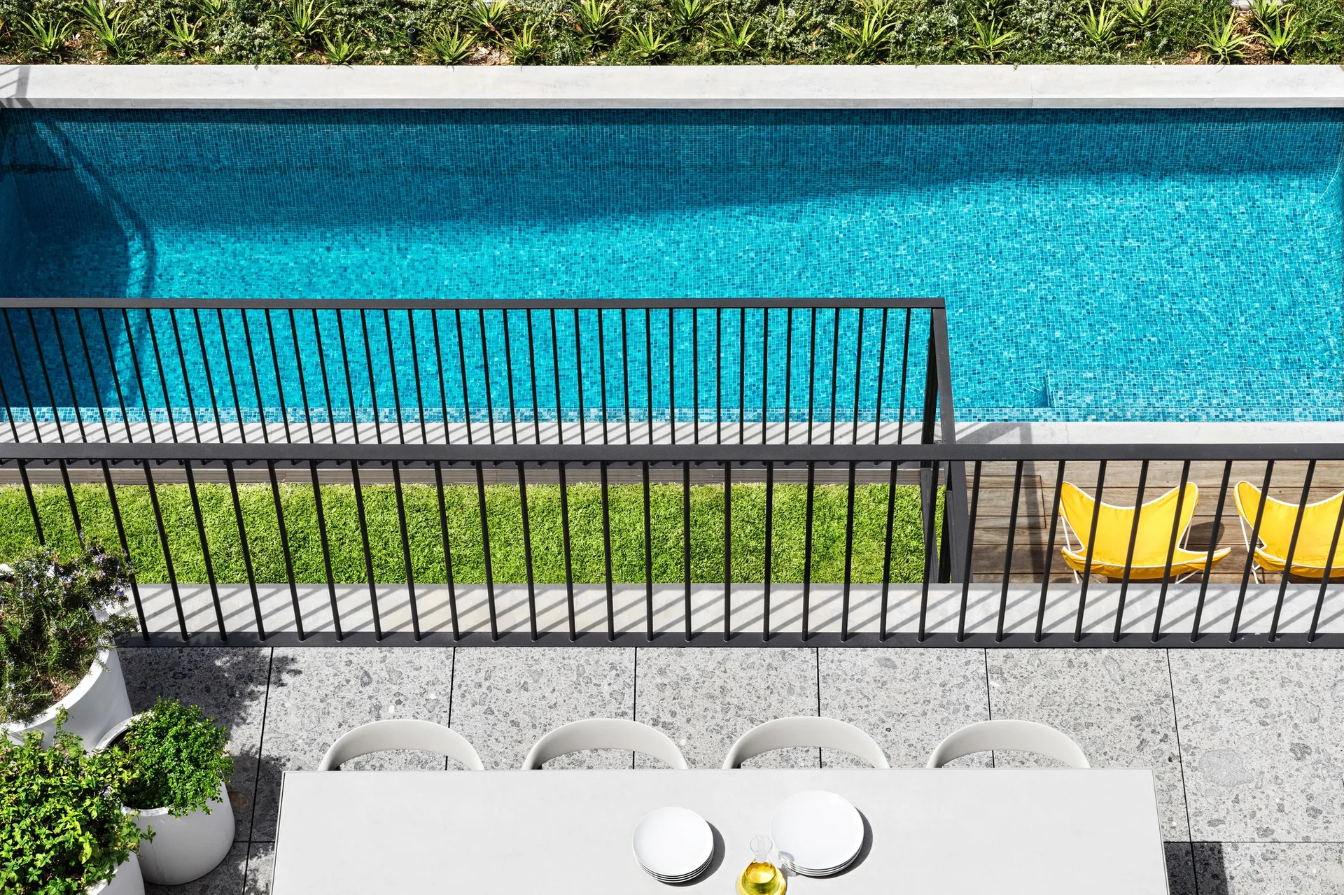 An overhead view of a grey stone patio with a white dining table next to a black metal railing overlooking a blue tiled swimming pool.