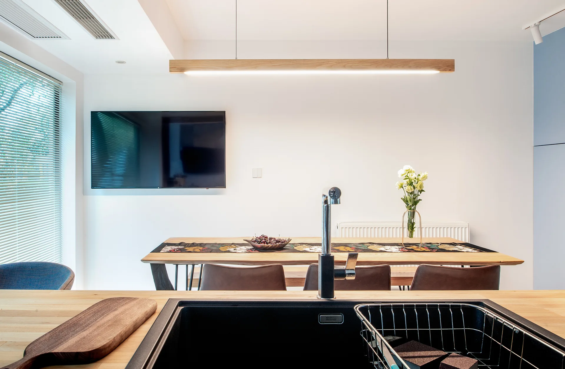 A black kitchen sink in the foreground looks out toward a wooden dining table with leather chairs and a long linear wooden pendant light.