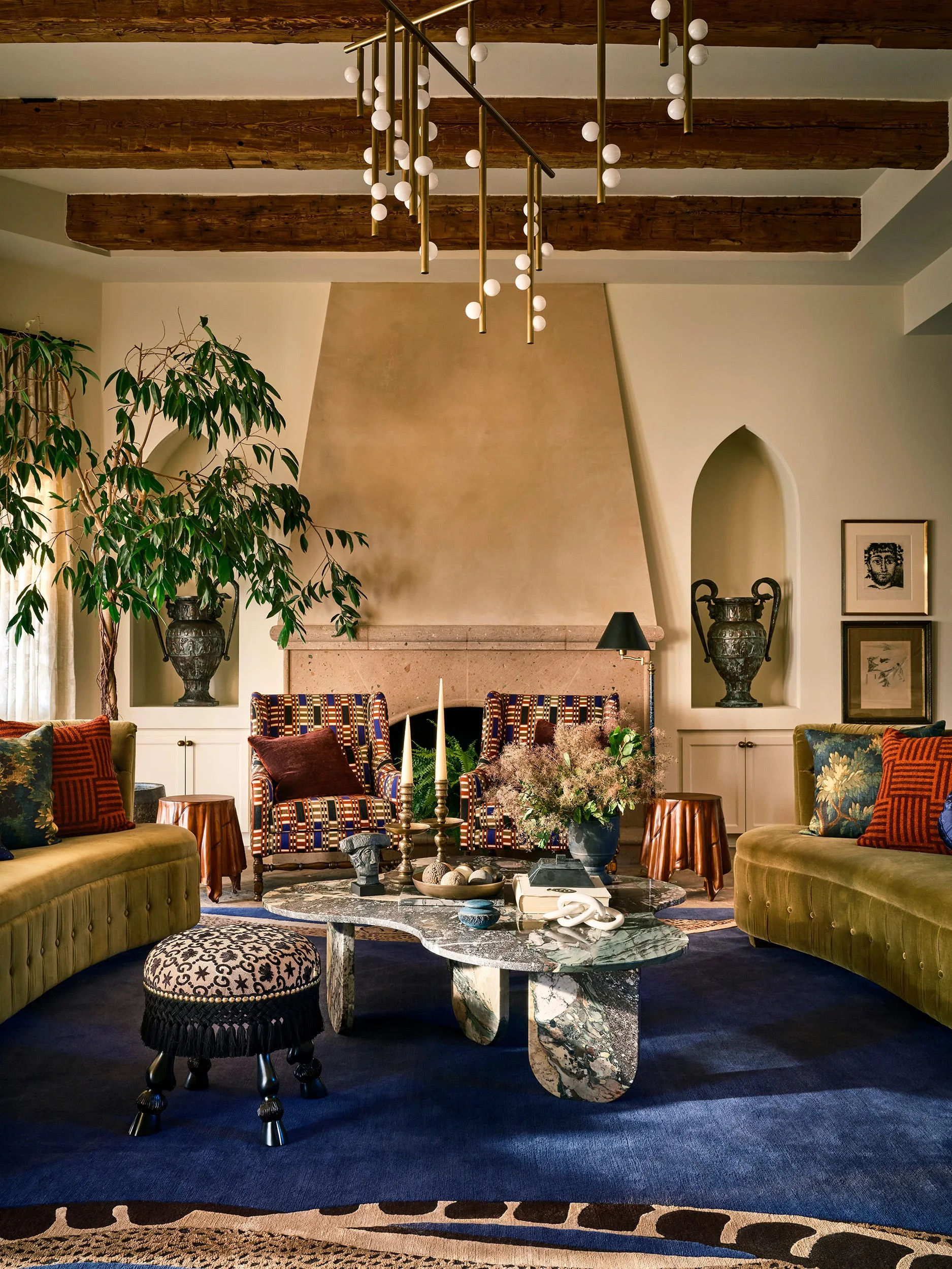 An eclectic living room features a stone fireplace, arched alcoves with urns, olive velvet sofas, and a marble coffee table on a navy rug.