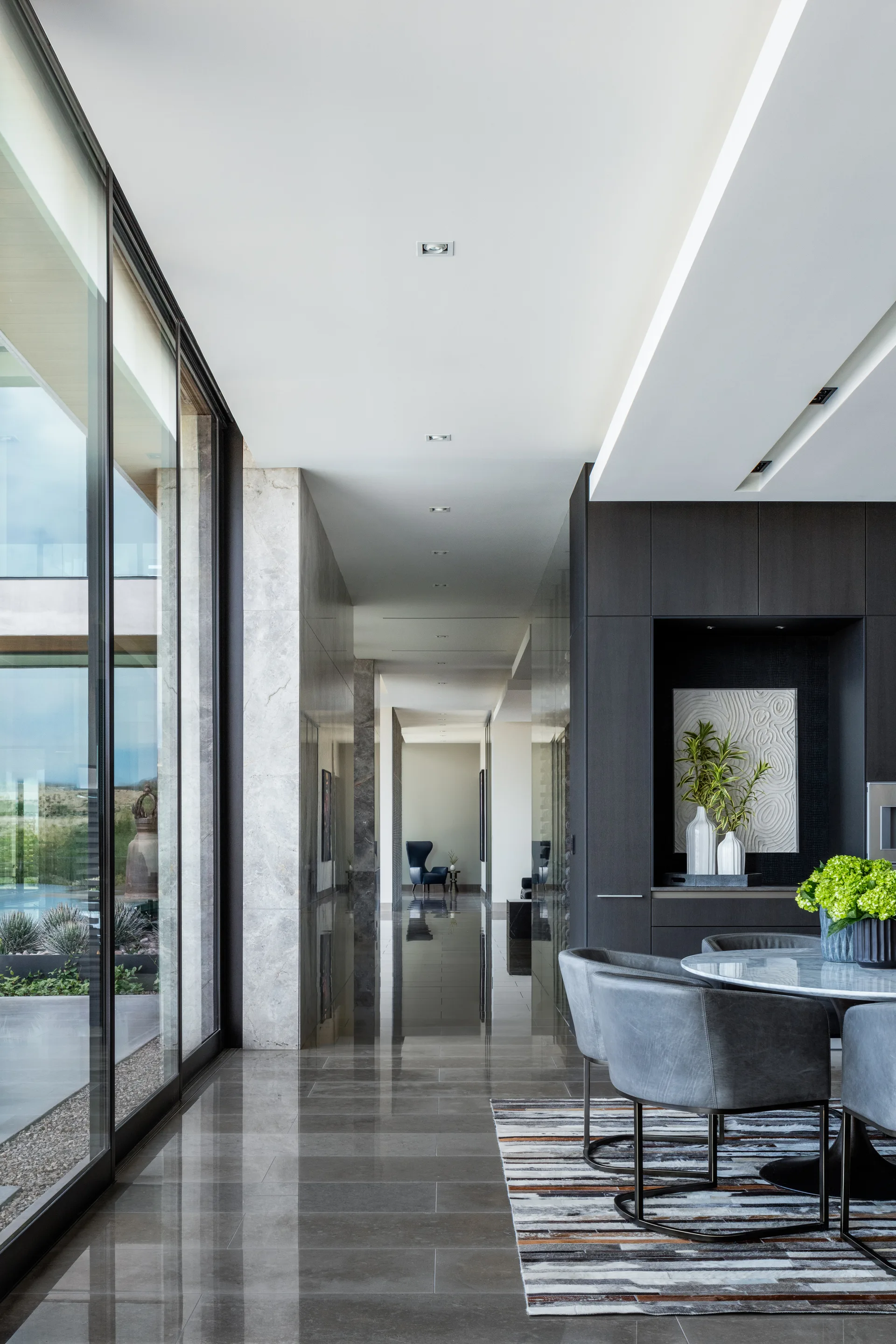 A long modern hallway with highly reflective grey stone flooring, a glass wall, and an adjacent dining area with dark cabinets.