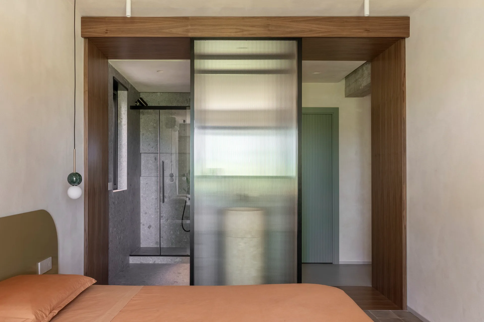 A bedroom with an orange bed looking through a wood-framed opening and fluted glass partition into a grey stone bathroom.