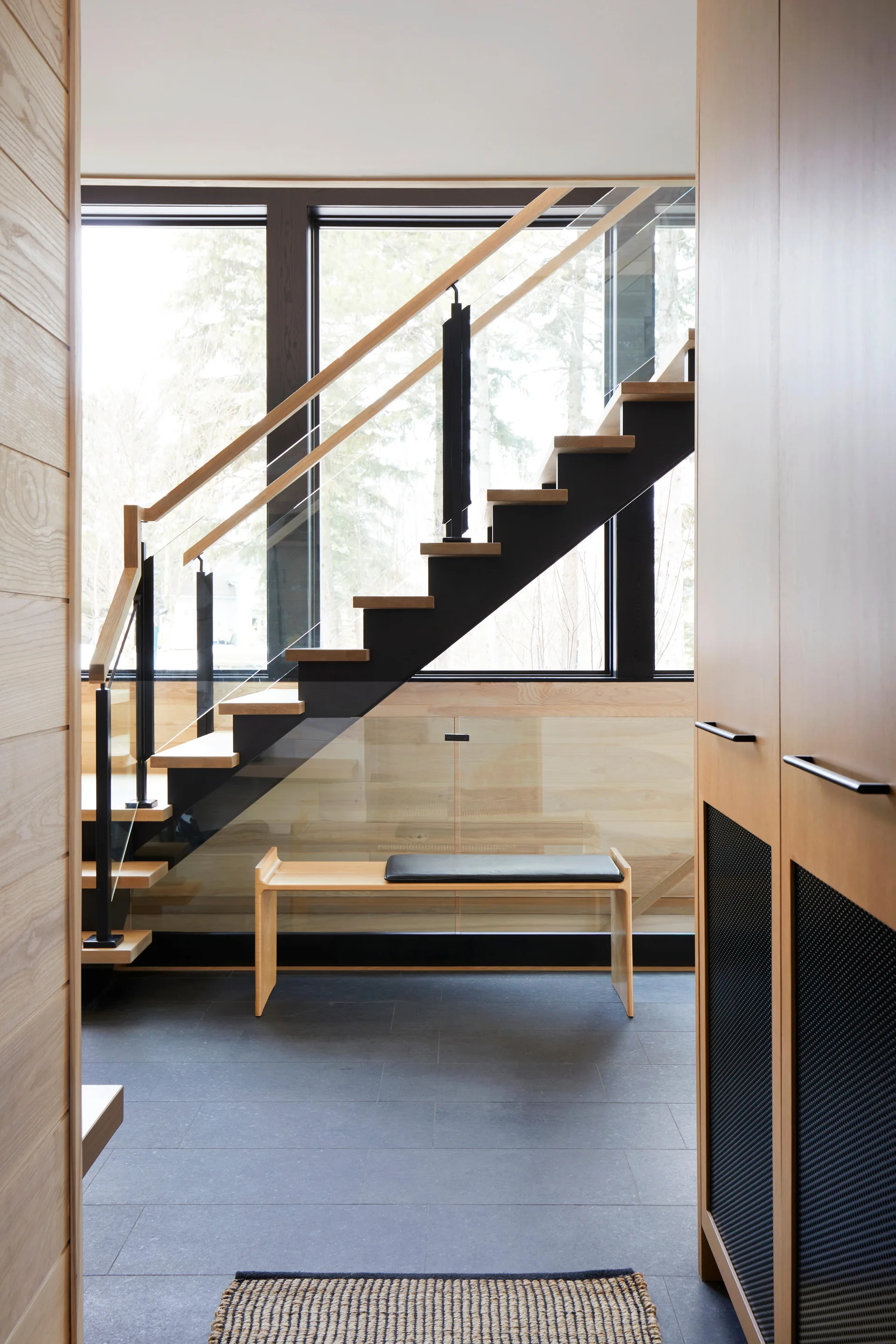 A modern entryway features an open-riser wooden staircase with a glass railing, a minimalist wood bench, and dark stone tile floors.
