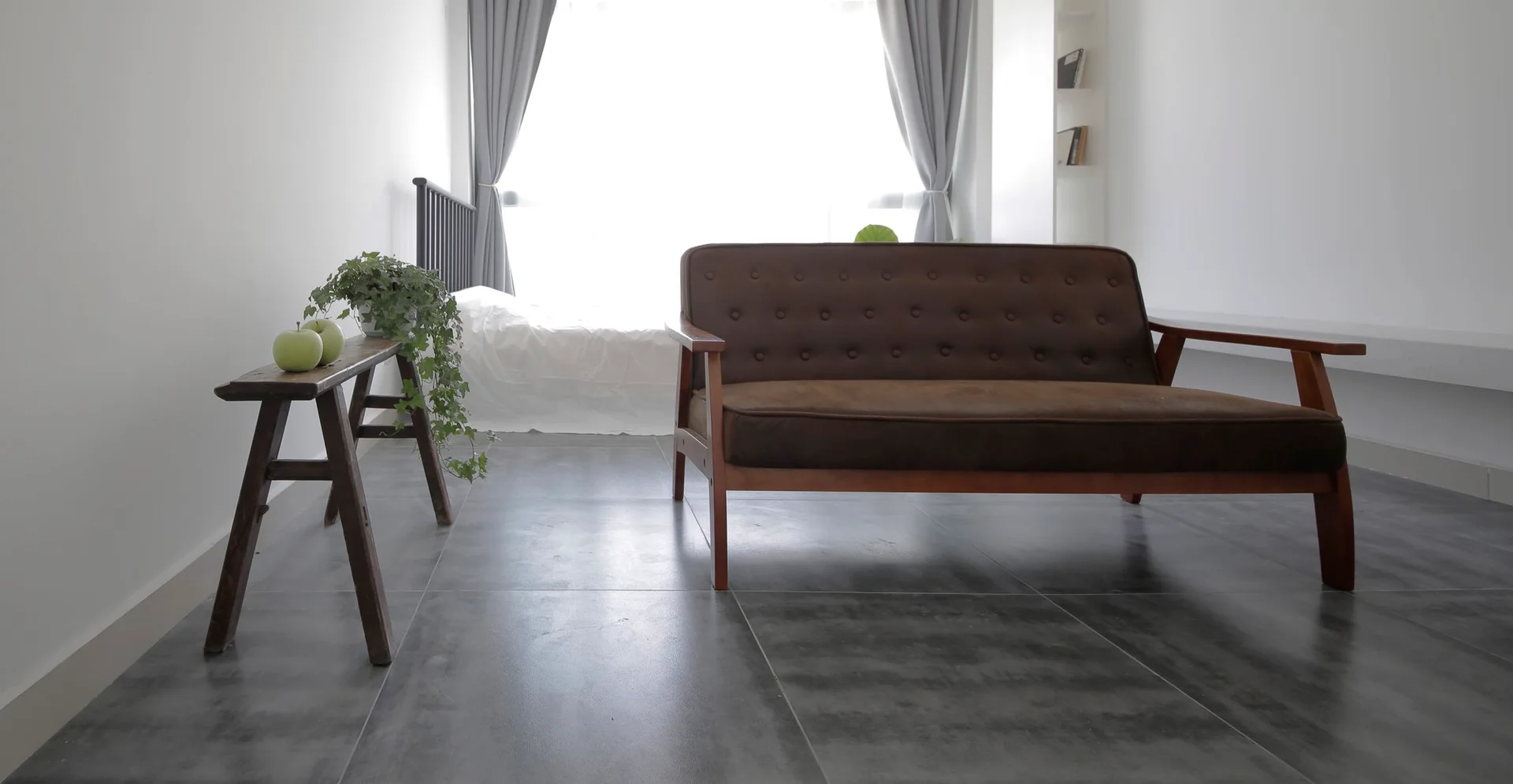 A minimalist room featuring a tufted brown sofa on large grey floor tiles next to a wooden bench holding a potted trailing ivy plant.