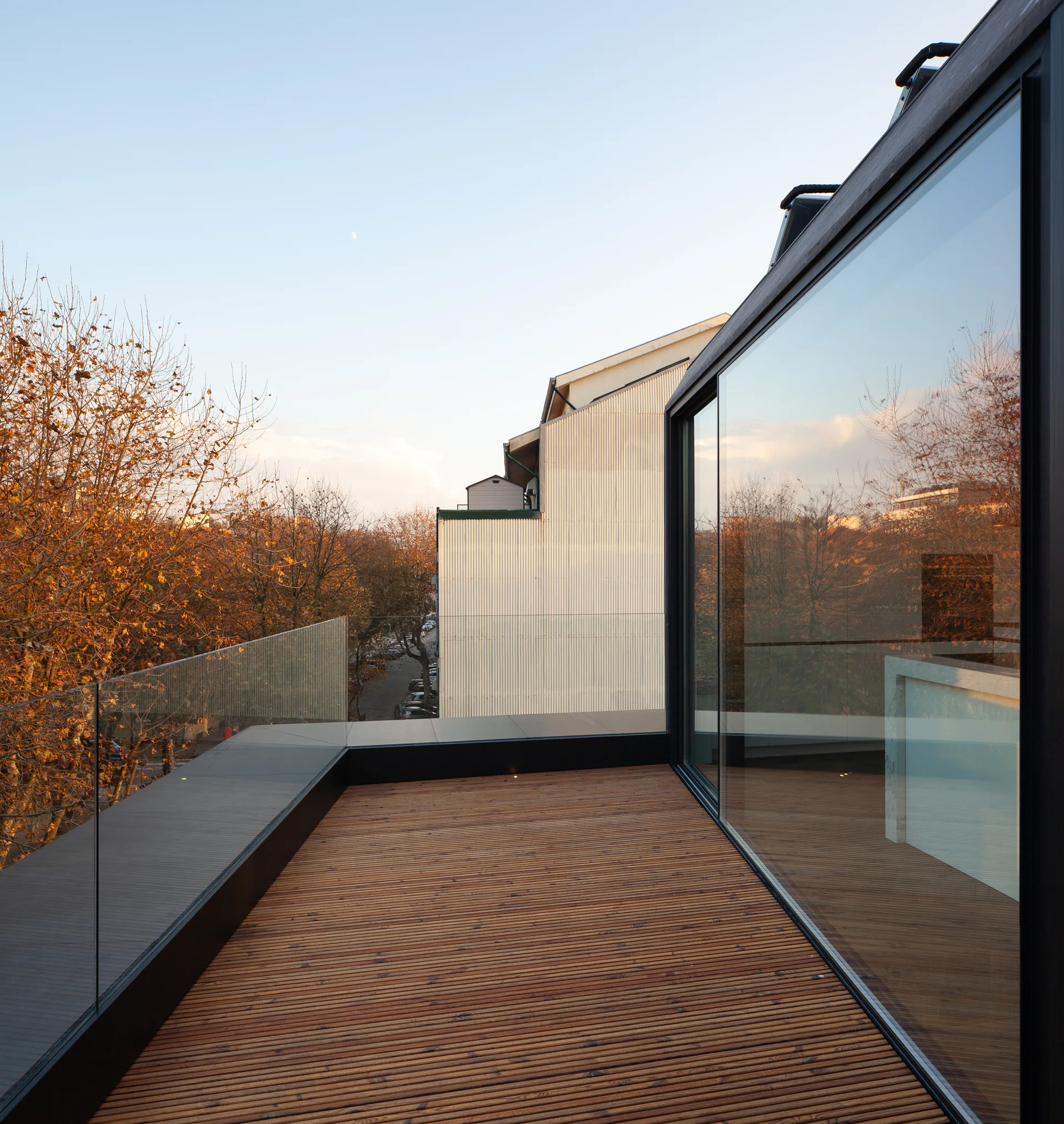 A modern balcony features a slatted wood deck floor, frameless glass railings, and a large black-framed sliding glass door overlooking autumn trees.