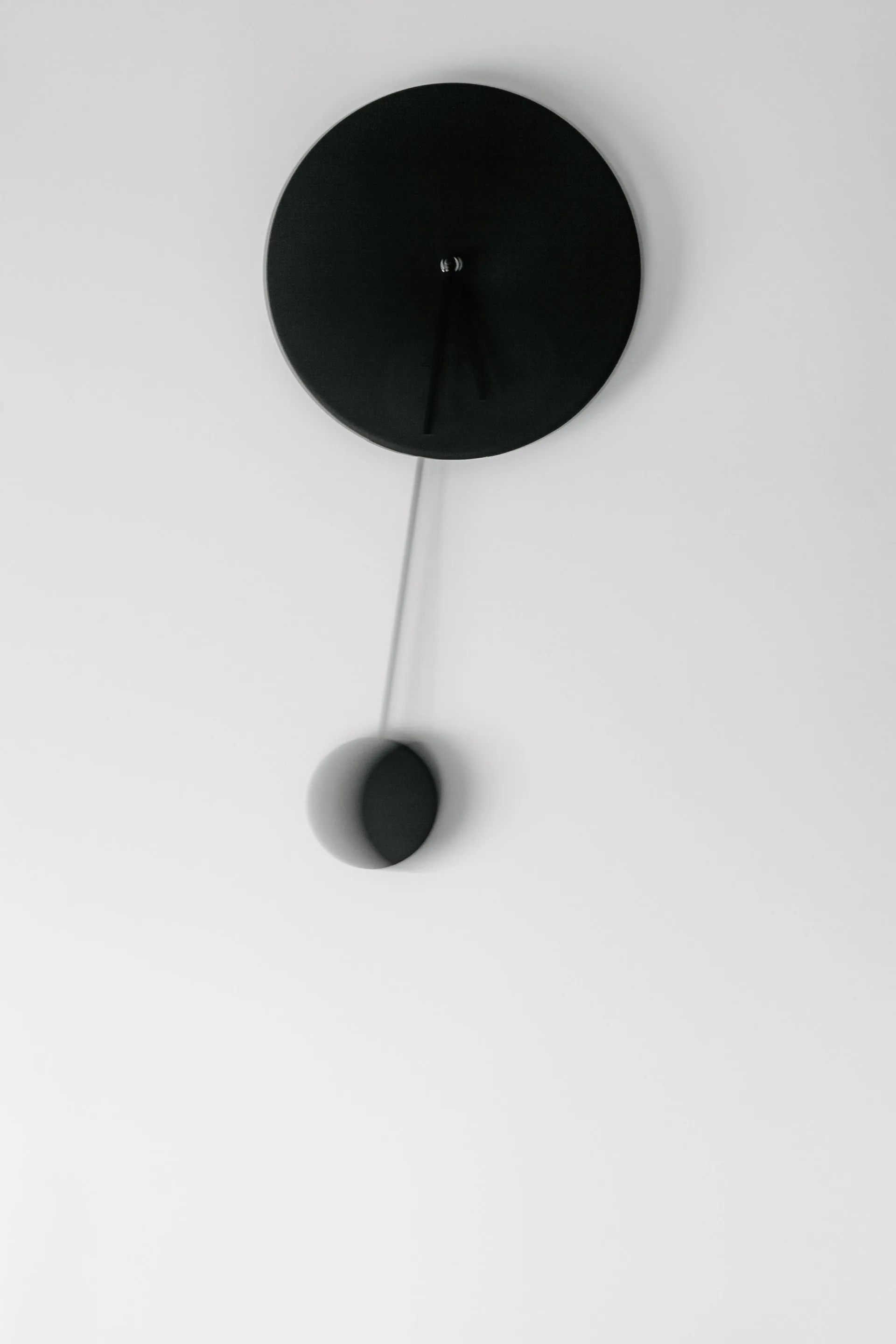A minimalist black circular wall clock with a swinging pendulum hangs against a solid matte white wall.