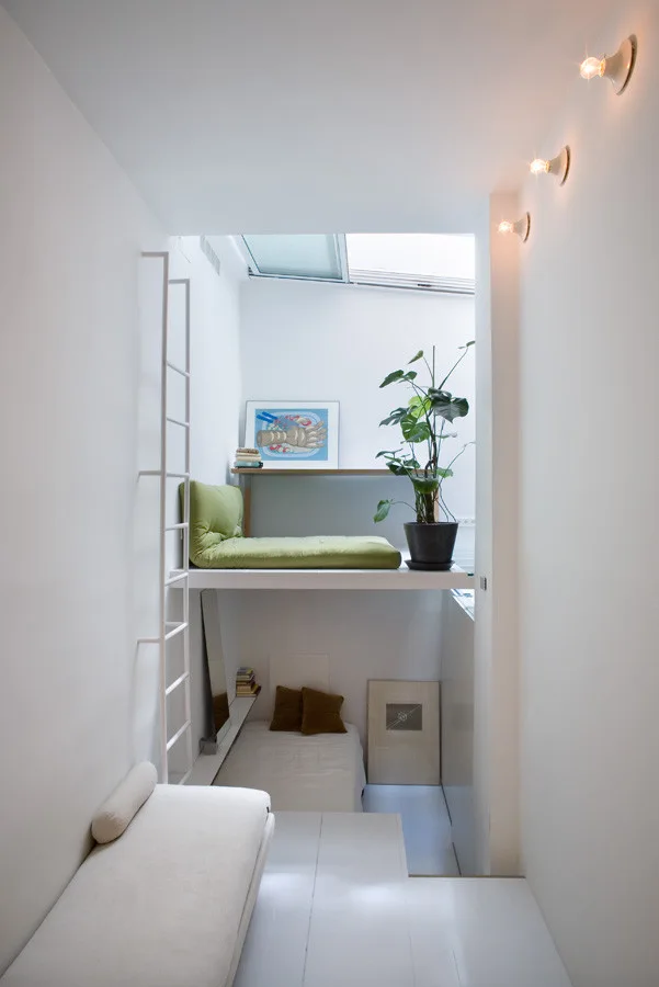 A narrow white bedroom with a lofted bed under a skylight, a lower sleeping area, and a white metal ladder against the wall.