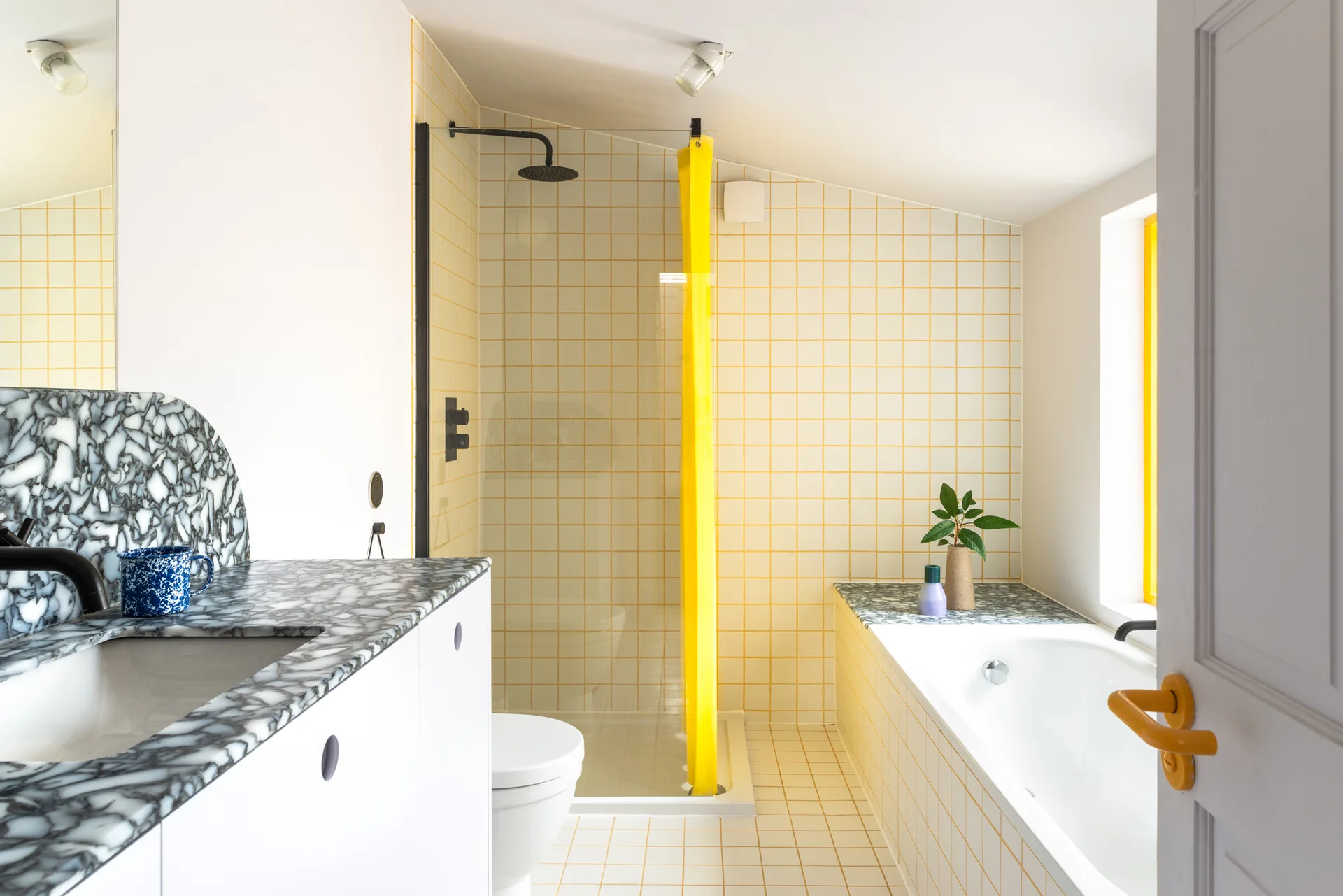A modern bathroom features yellow square grid wall tiles, a built-in white bathtub, and a vanity with a highly figured black and white marble countertop.