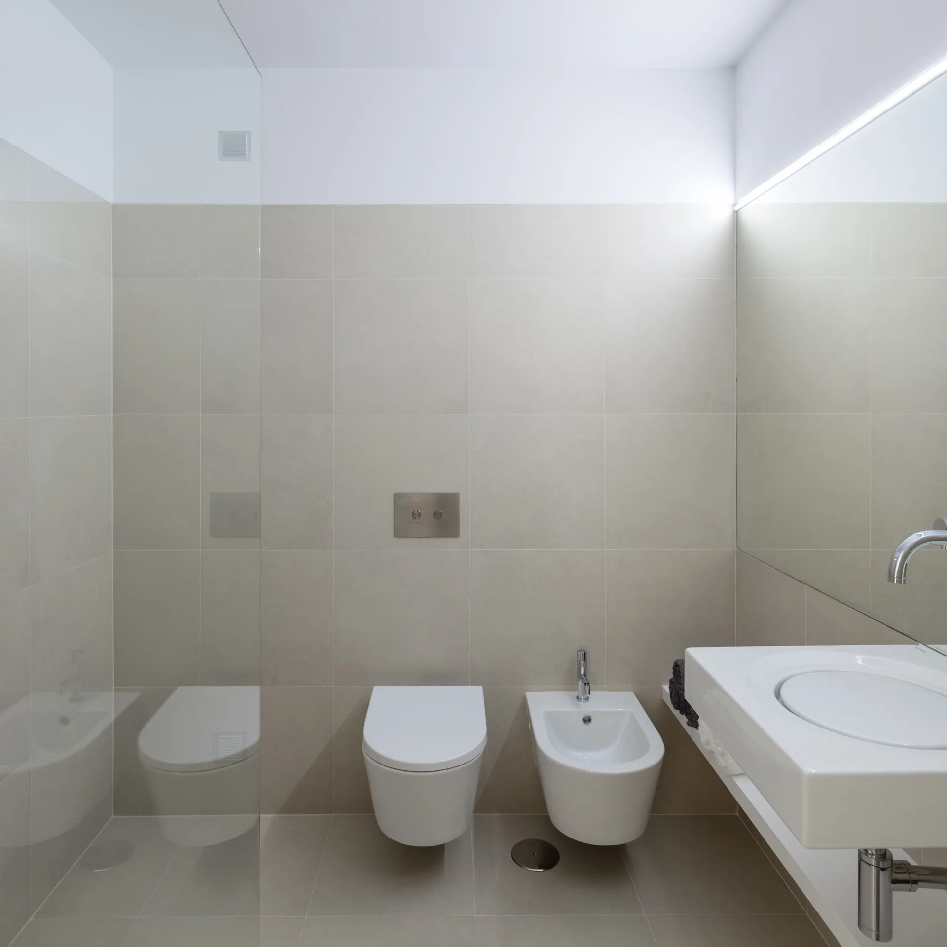 A modern minimalist bathroom featuring a wall-mounted toilet and bidet next to a white sink basin and large beige wall tiles.