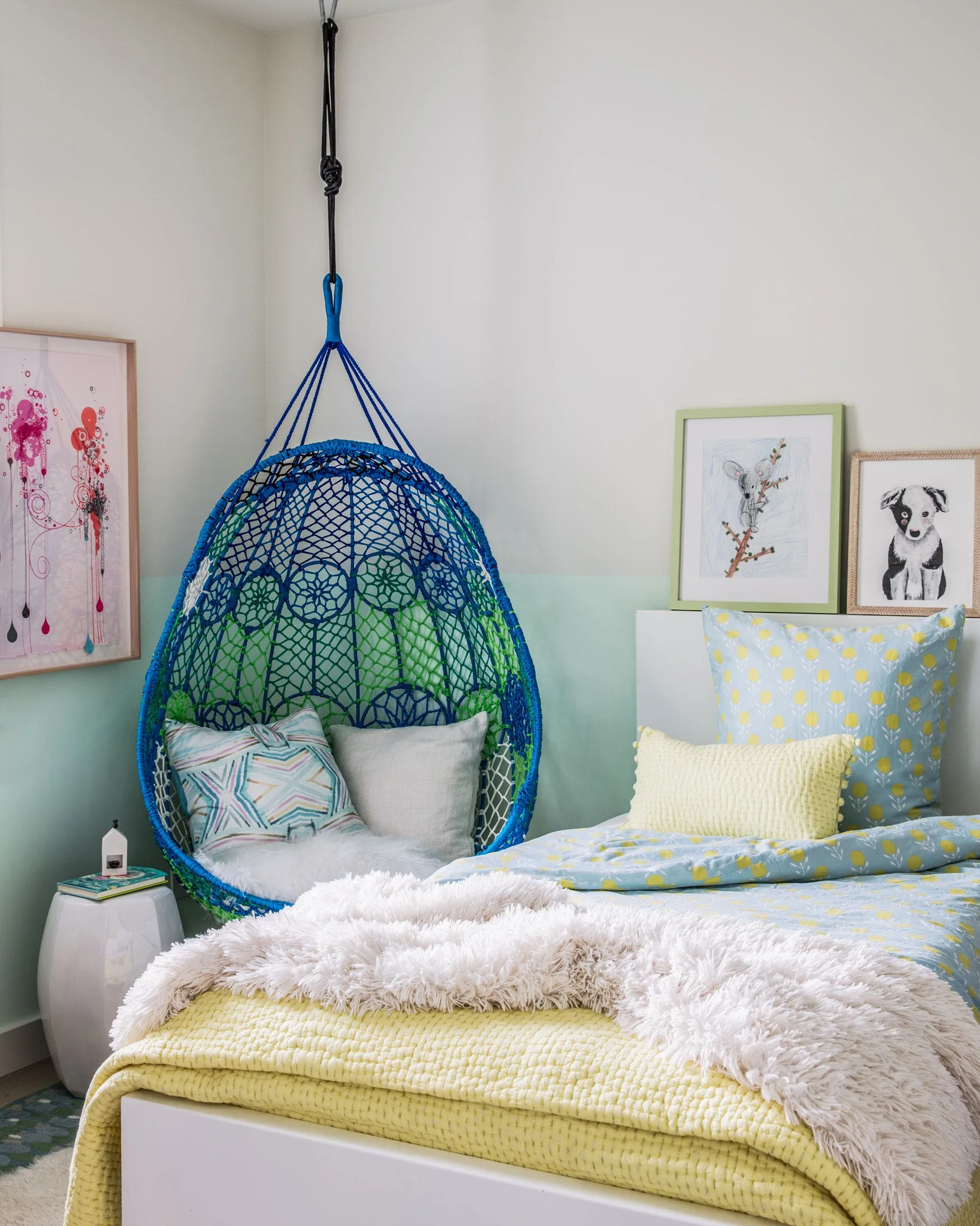 A children's bedroom featuring a blue and green hanging macrame chair in a corner next to a bed with yellow and patterned bedding.